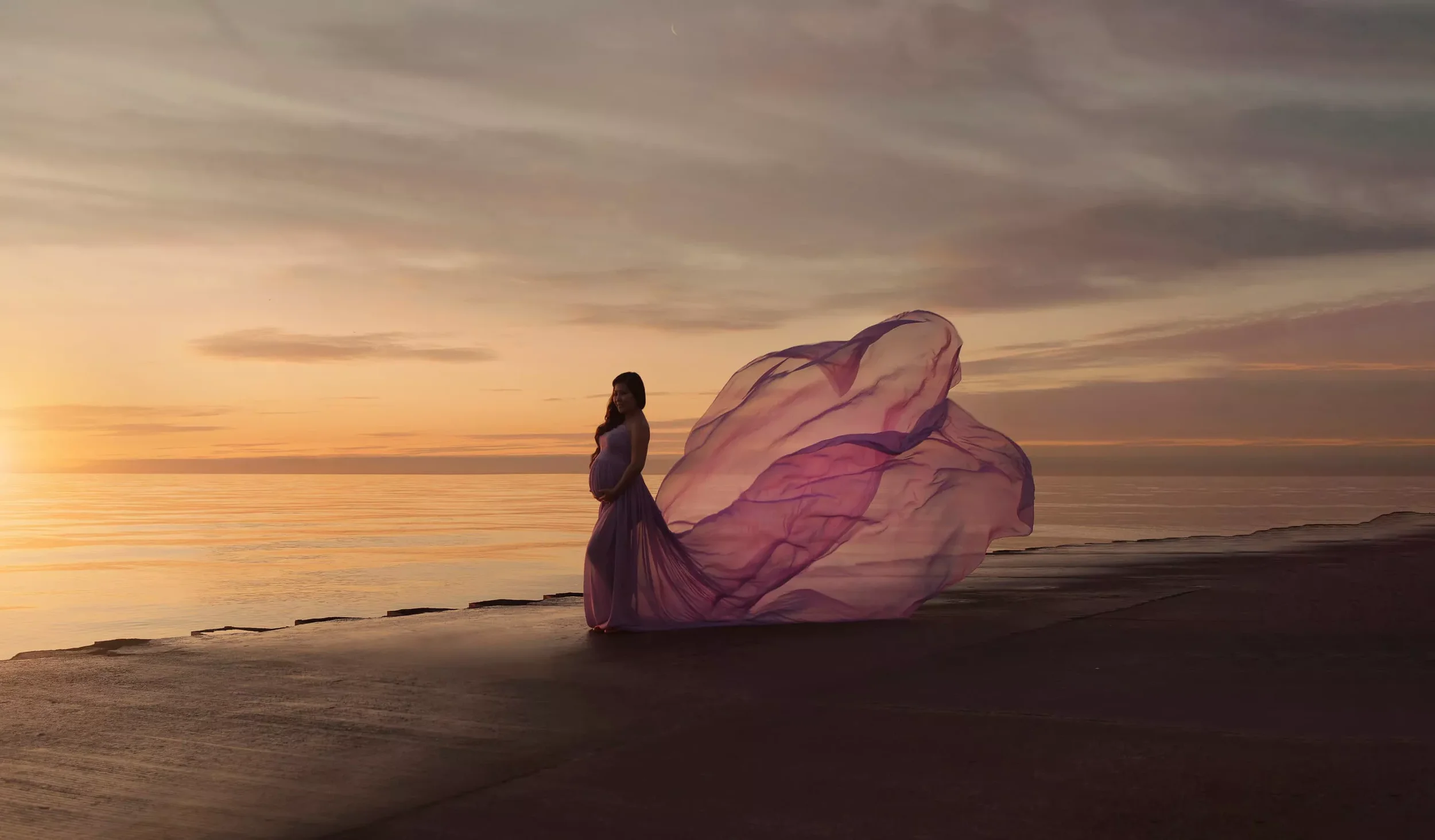 A pregnant woman in a flowing pink dress standing on a beach at sunset, with large pink and purple fabric extending from her back.