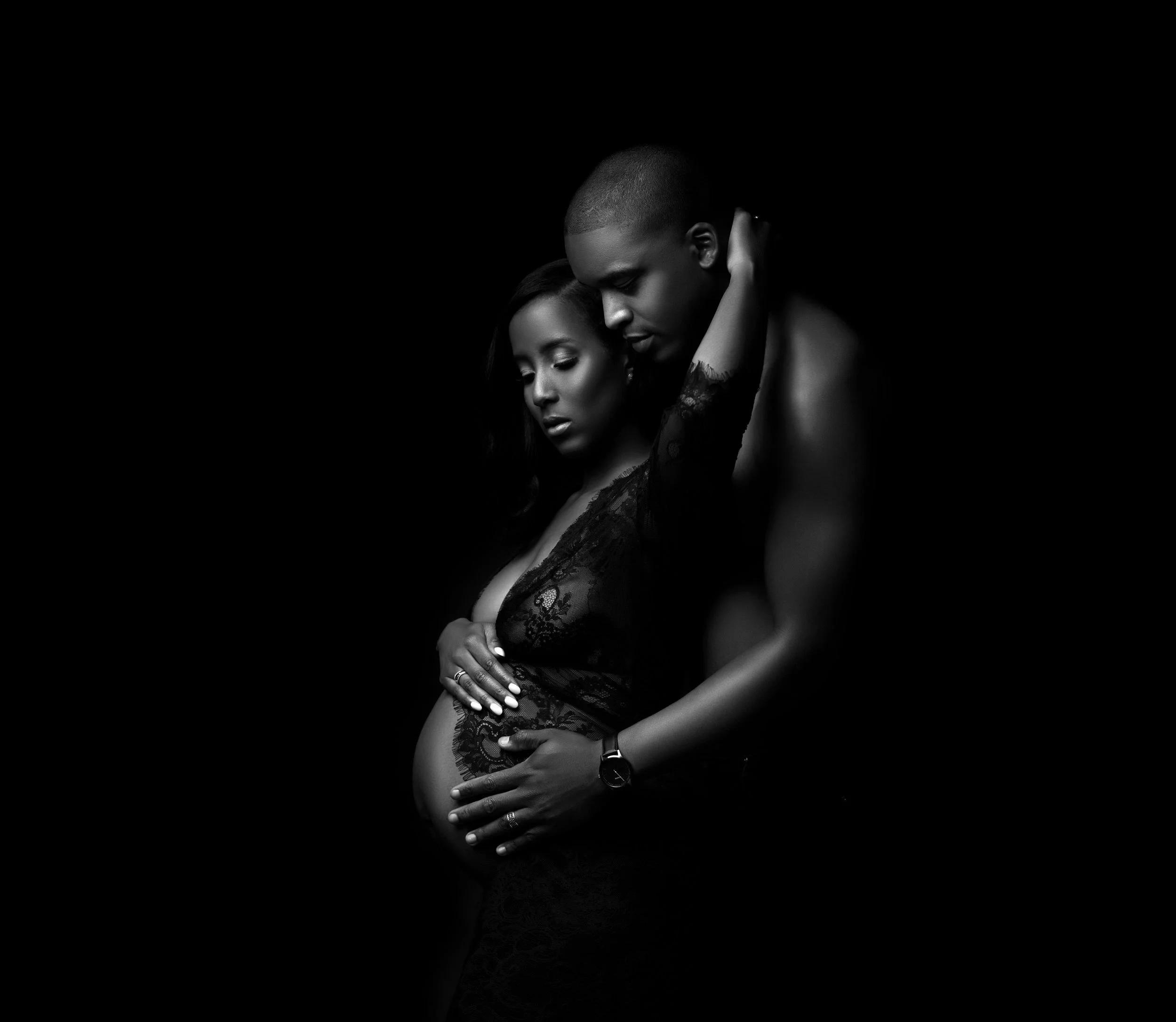 A black-and-white photograph of an intimate pose between a man and woman, with their foreheads touching and eyes closed. The woman has long dark hair and is wearing lace clothing, while the man has a shaved head and muscular build, with his arm aroun