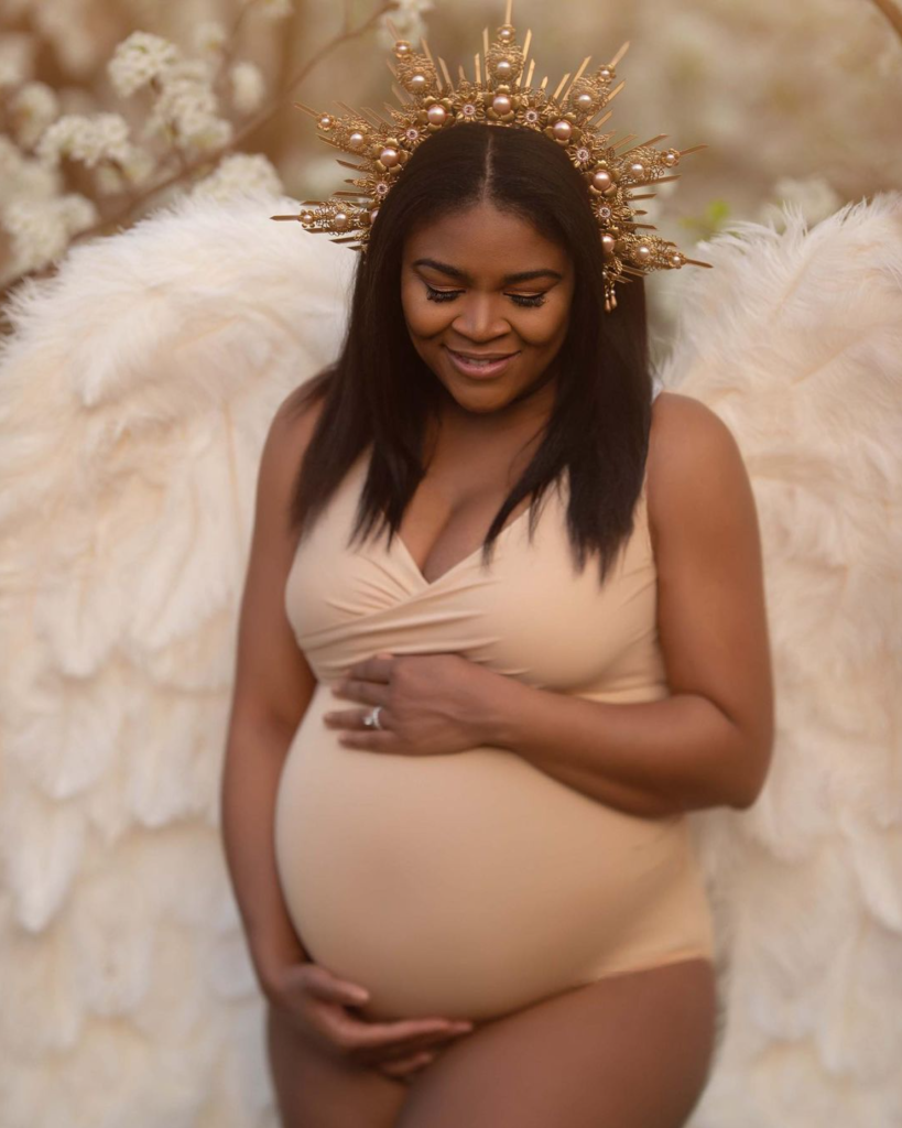 Pregnant woman with large angel wings, wearing a beige top and a decorative golden crown with pearls, smiling softly with her hands on her belly, standing in front of a soft floral background.