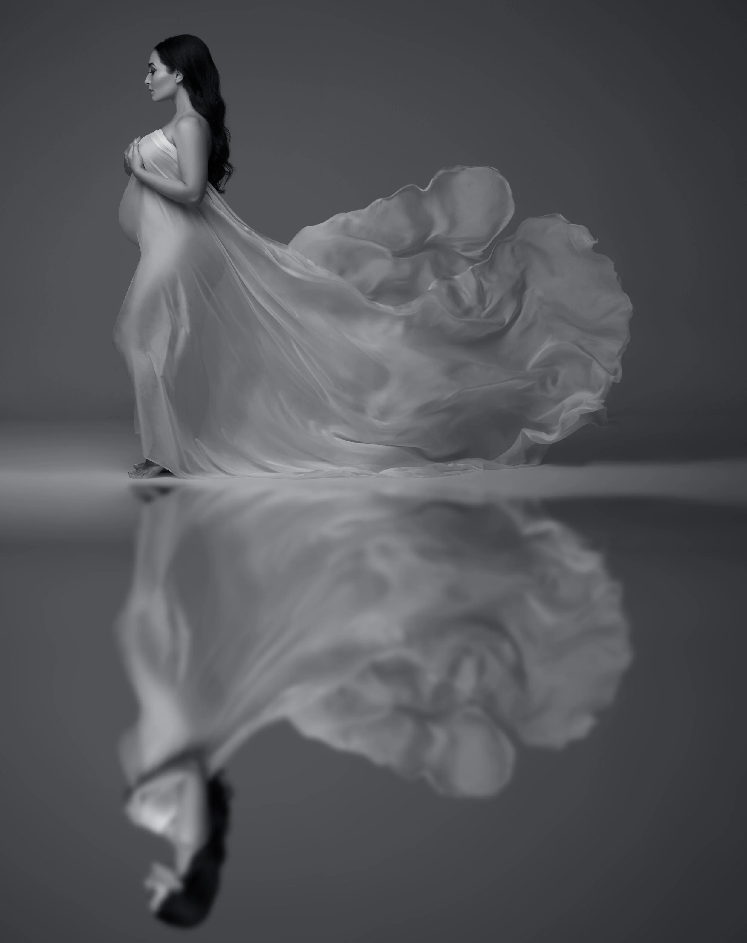 Elegant B&W maternity photography with reflection of a pregnant woman in flowing silk against grey background.