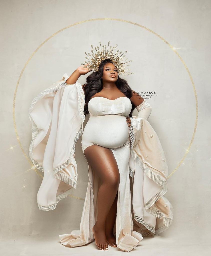 A woman with dark, wavy hair wearing a white strapless gown and a crown standing against a neutral background. She is posing with her left hand on her hip and her right hand adjusting her crown.
