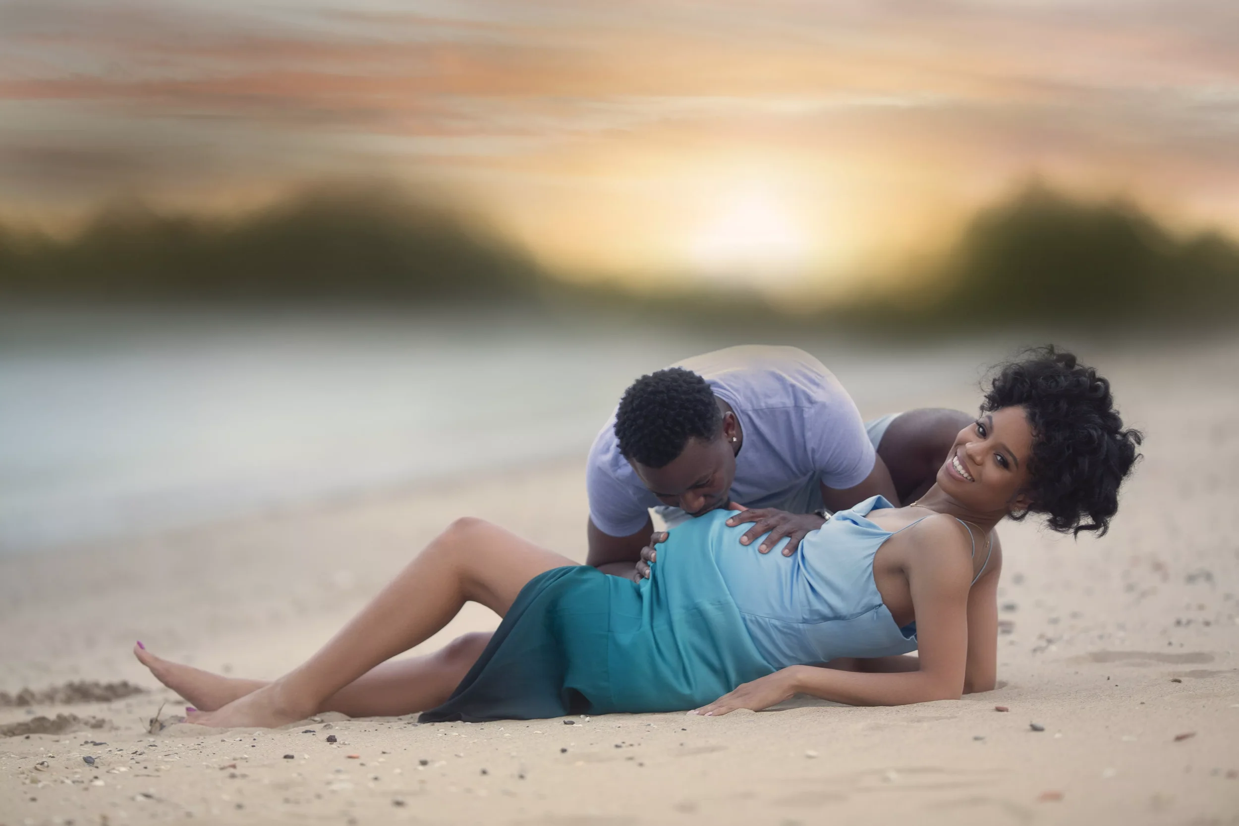 A couple having fun on the beach, with the man leaning over the woman who is lying on the sand, both smiling and enjoying their time together at sunset.