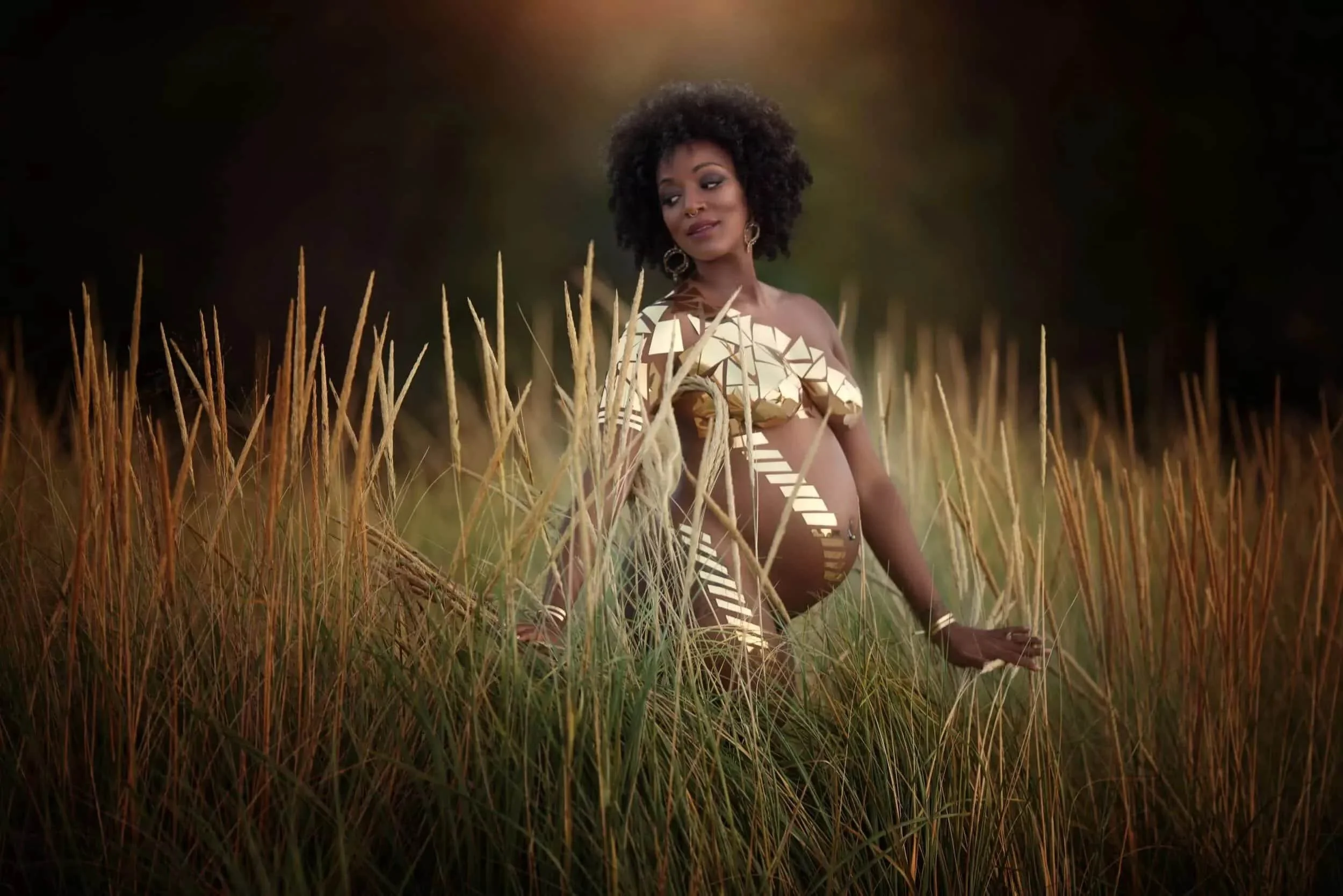 A woman with curly hair and dark skin standing in a field of tall grass, wearing a patterned outfit with cutout details, and looking to the side with a serene expression.