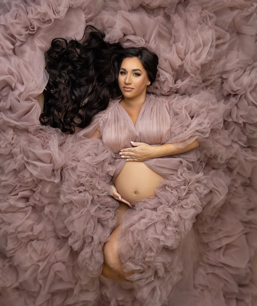 A pregnant woman with dark hair lying on a bed of pink ruffled fabric, gently touching her belly and looking at the camera.