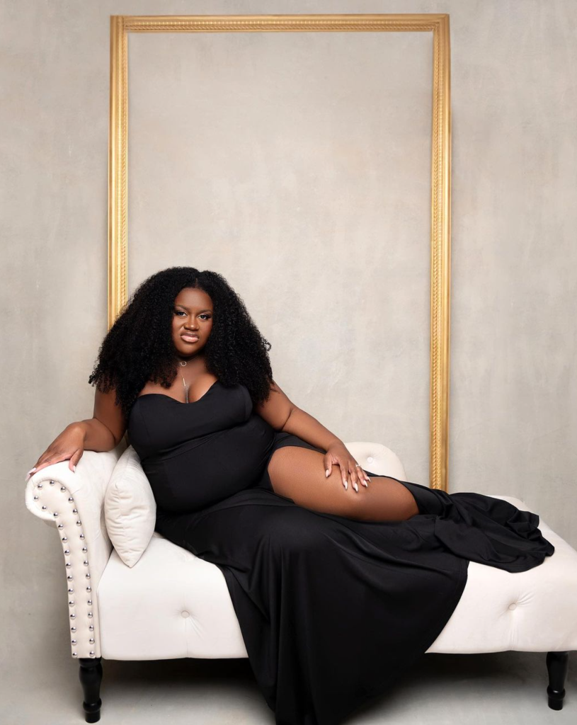 A plus-sized woman with curly black hair lounging on a white chaise lounge, wearing a black dress and thigh-high stockings, in front of a neutral background with an empty gold picture frame.