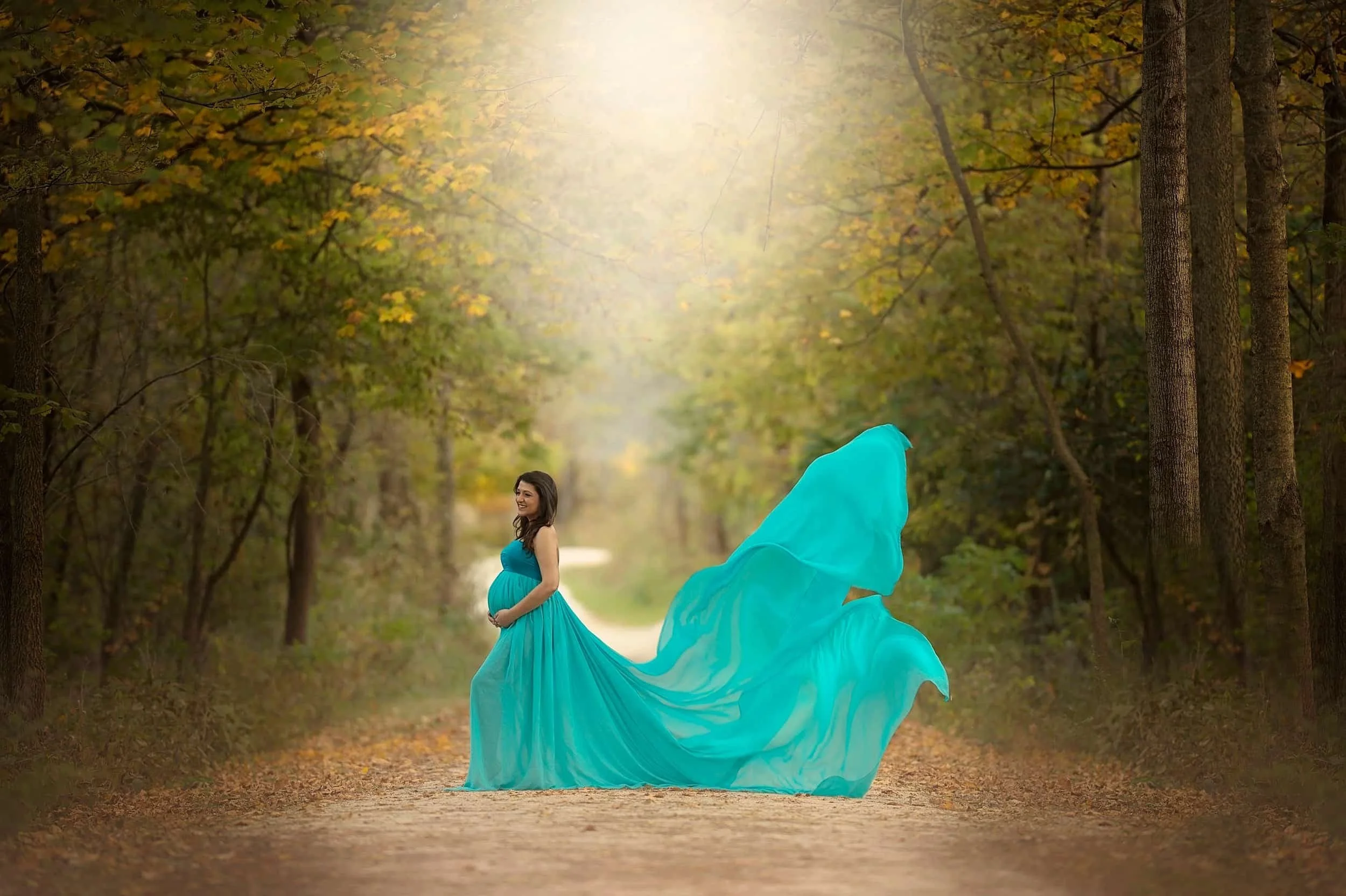 Chicago pregnant woman in a flowing turquoise gown standing on a dirt path in an autumn forest
