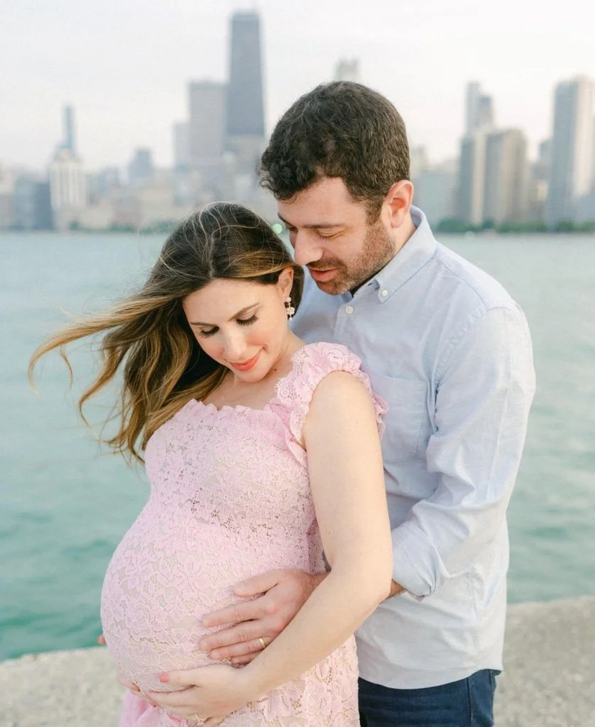 A couple embraces by the water; the woman is pregnant in pink, city skyline behind—a Chicago maternity photographer's dream.