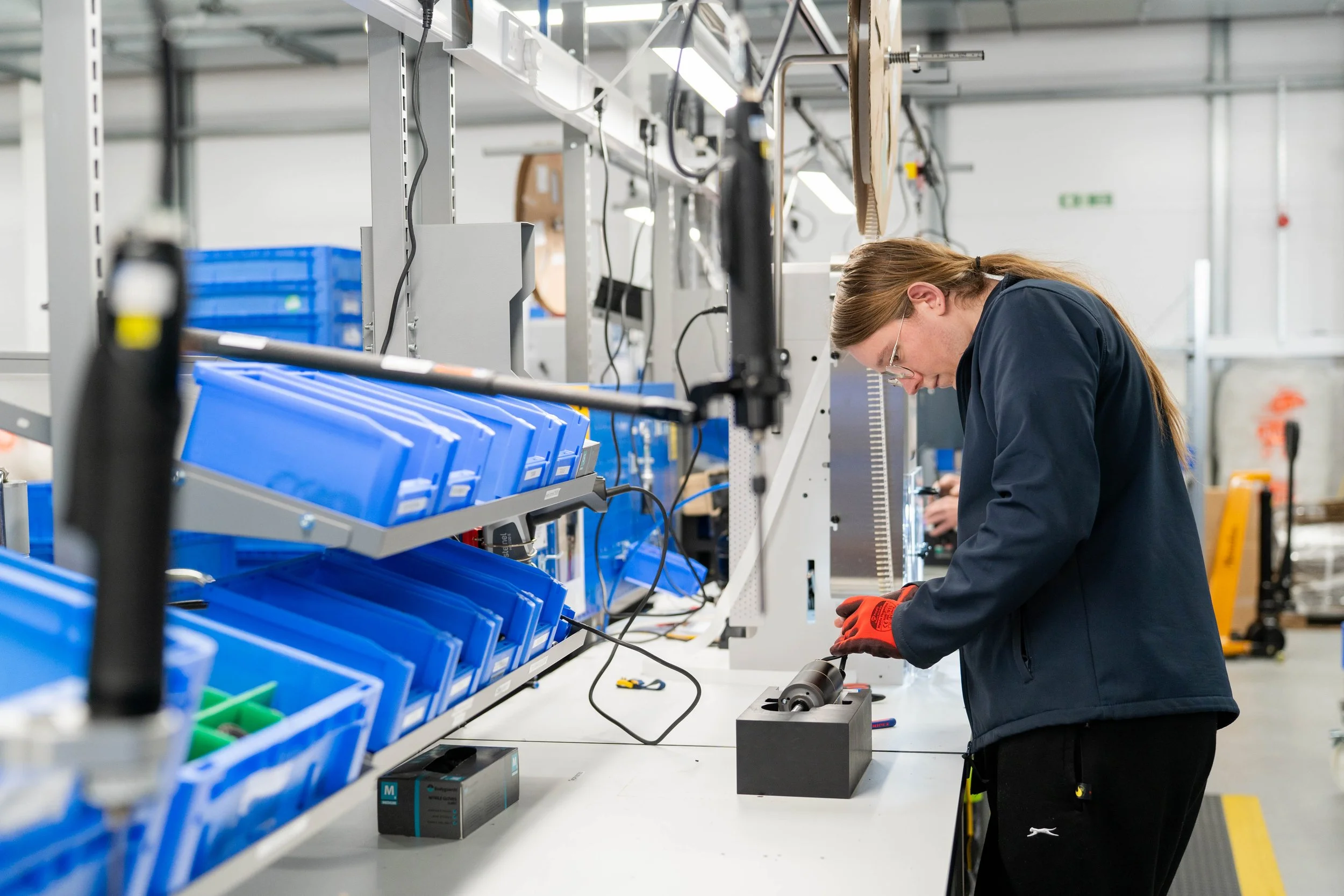 A woman wearing safety glasses and gloves works at a factory bench, assembling a mechanical component using hand tools. Blue storage bins and industrial equipment line the workstation, shown here through industrial photography.