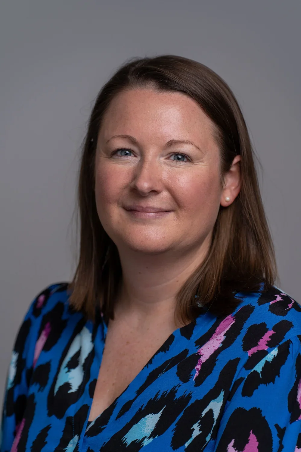 Corporate headshot of a businesswoman in a photography studio in London. The subject has shoulder-length brown hair and is wearing a blue and pink patterned blouse.
