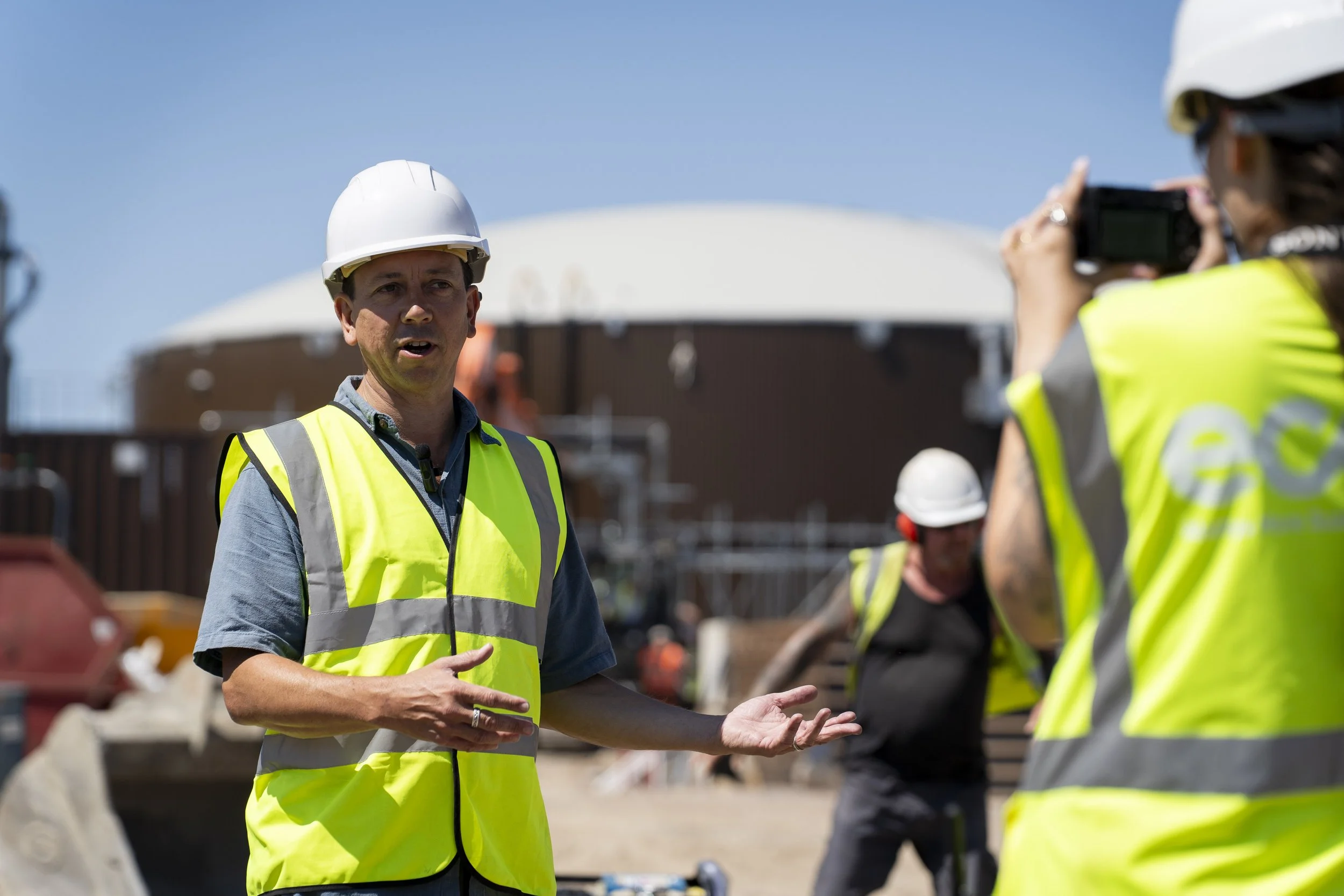 A man wearing a hard hat and high-visibility vest speaks while gesturing on an active construction site. Other workers and large industrial tanks are visible behind him, captured as part of a construction photography shoot.
