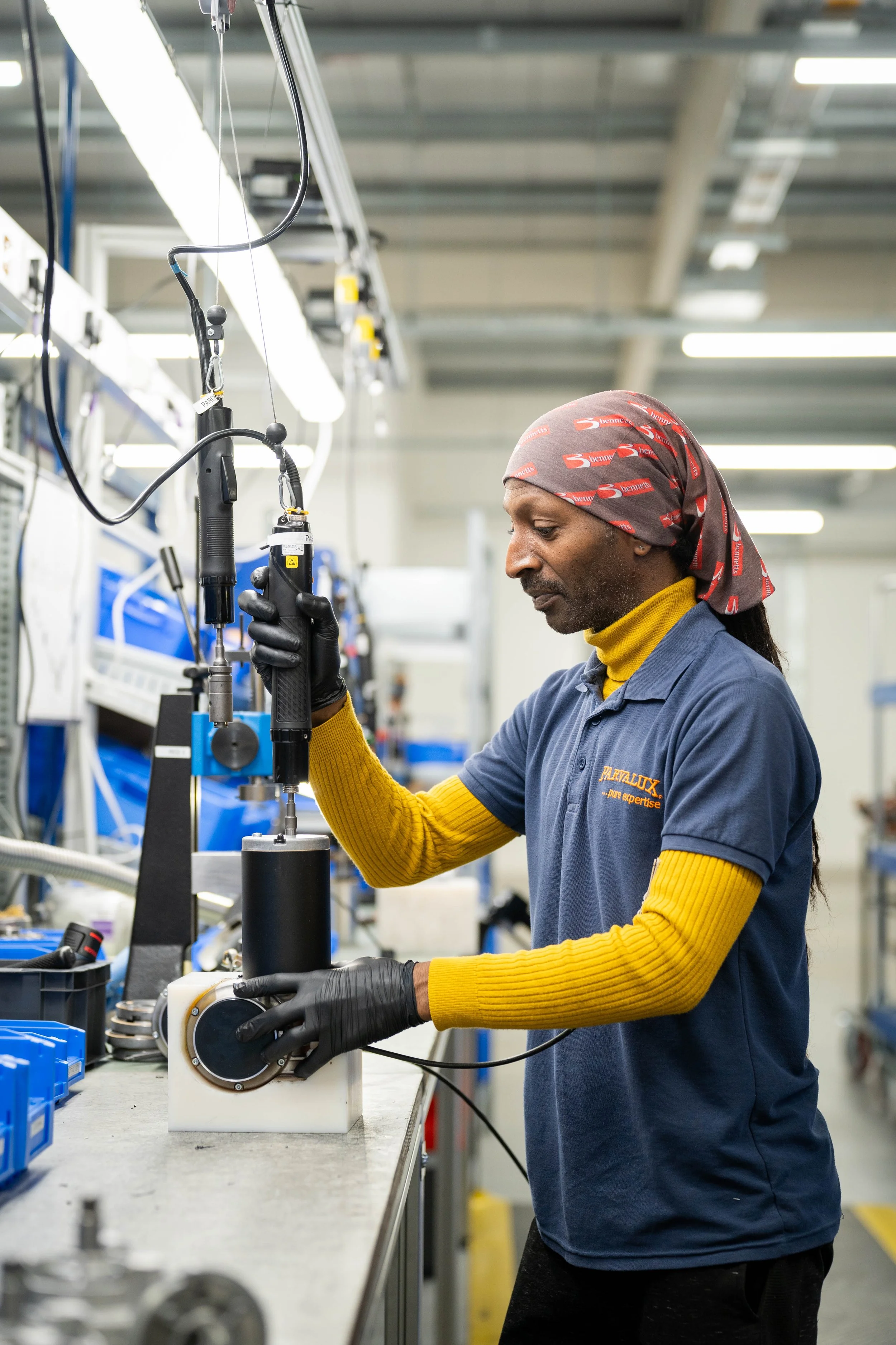 A factory worker uses a suspended powered tool to assemble a motor unit at a production bench. The image shows industrial photography capturing precision assembly work inside a manufacturing environment.