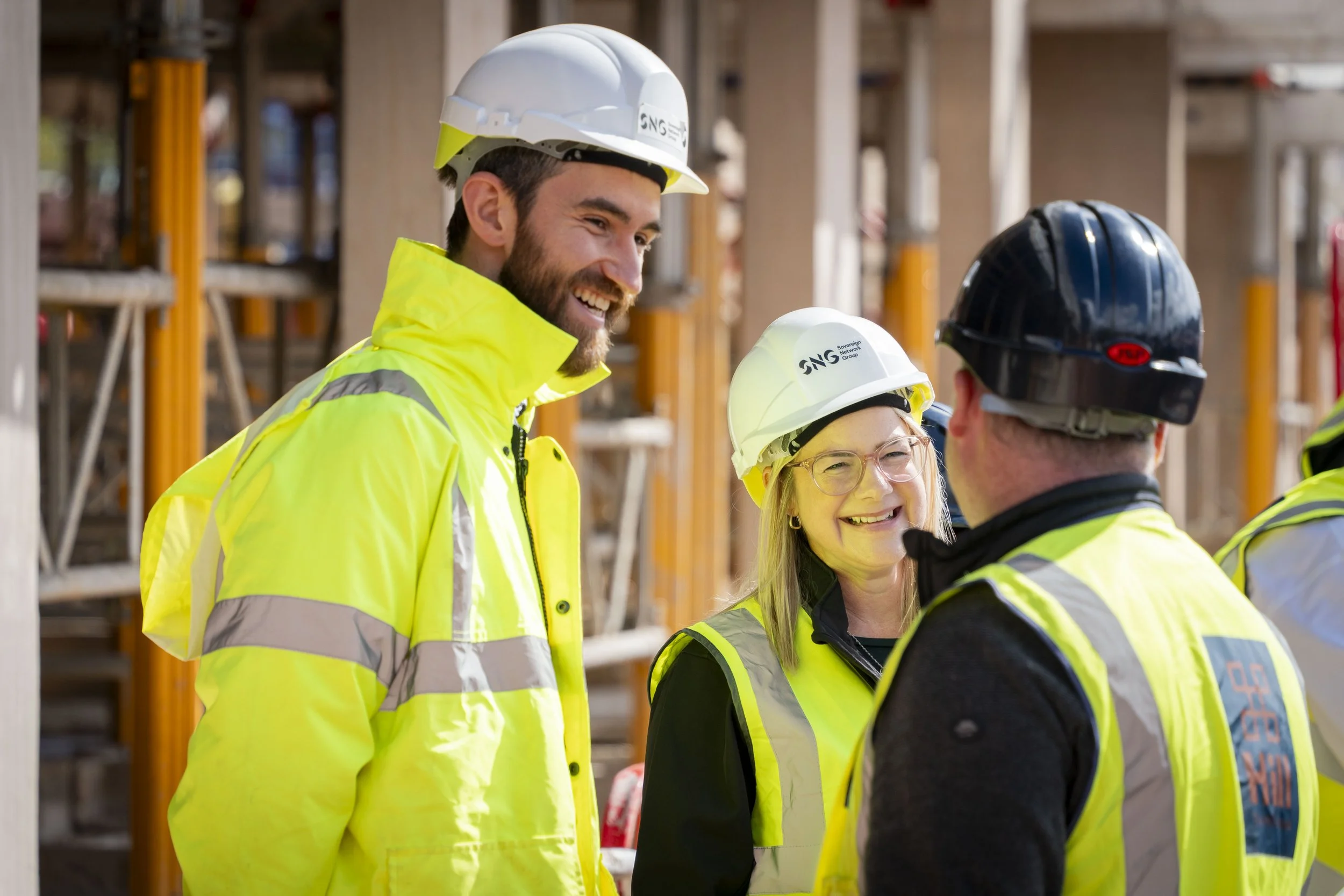 Three construction workers wearing hard hats and high-visibility jackets talk together on an active building site. The image shows a candid moment often captured in construction photography, focusing on real interactions between workers.