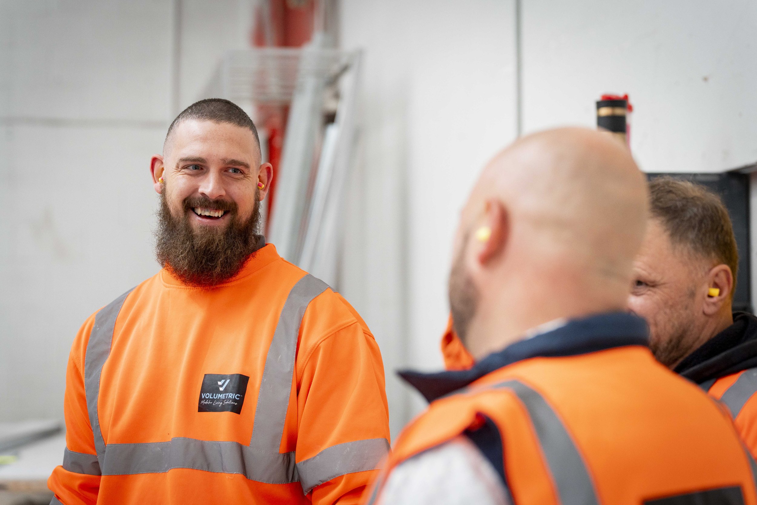 A construction worker in high-visibility orange clothing smiles while talking with colleagues on an indoor work site. The image reflects commercial photography focused on people and communication within a construction environment.
