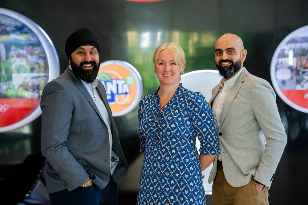 Team photography of three business professionals, one female and two male. They are standing in an office lobby in front of a wall emblazoned with the logos of soft drinks such as Fanta.