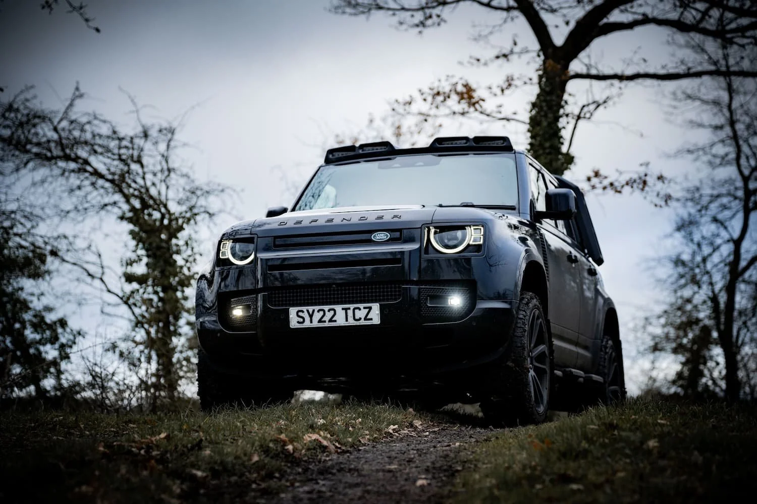 Dark green Landrover Defender is parked on top of a grassy hillside. The photograph is taken from down the hillside looking up at the vehicle which is parked below a tree and has its headlights on.