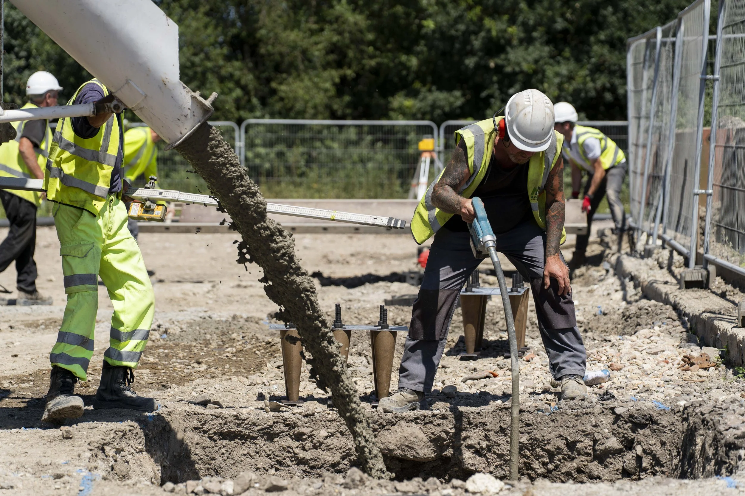 Construction workers in hard hats and high-visibility clothing pour wet concrete into a trench using a chute and vibrating tool. The scene is captured as construction photography, showing groundworks and on-site building activity in progress..