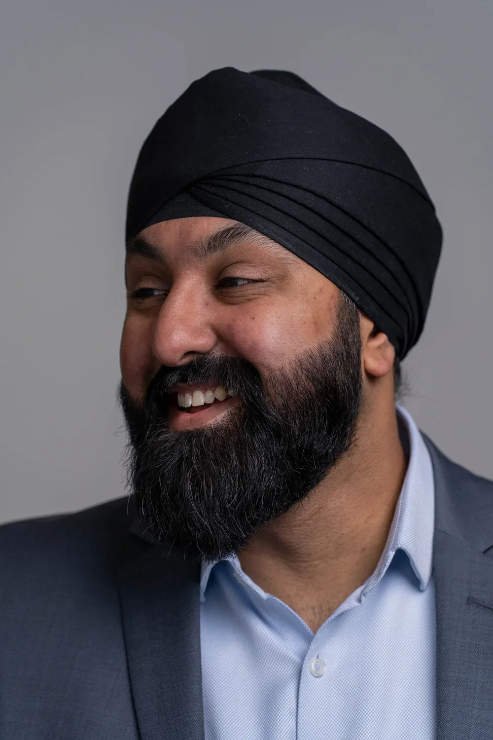 Friendly corporate headshot photography of man wearing a black turban. He is smiling in a relaxed manner and is looking slightly away from the camera to the left.