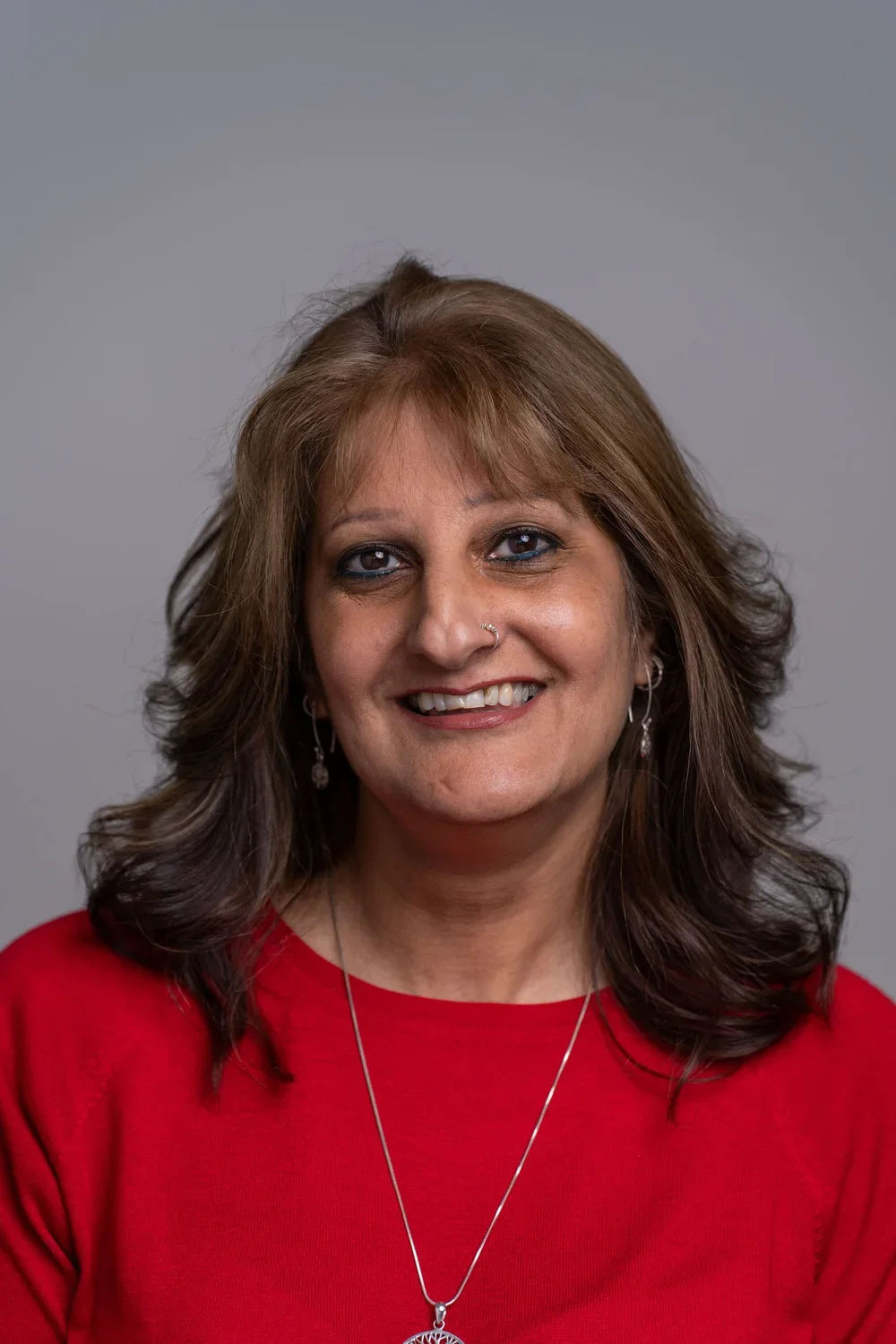 Business portrait of an Indian woman in London. She has shoulder-length brown hair and is wearing a bright red jumper and silver necklace.