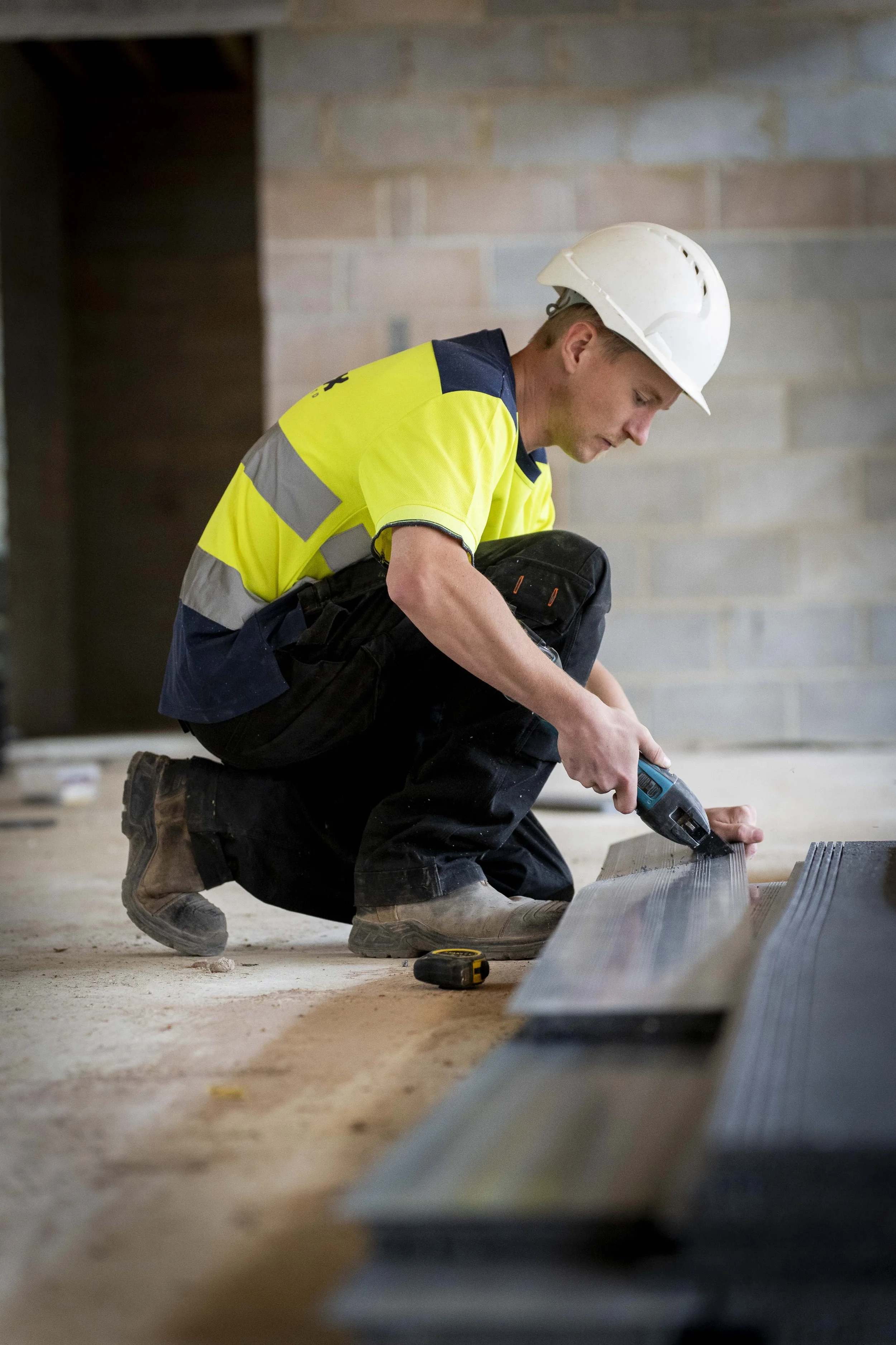 A construction worker wearing a hard hat and high-visibility clothing kneels on the floor while fixing metal channels with a power drill. The image shows commercial photography focused on hands-on construction work inside an unfinished building.