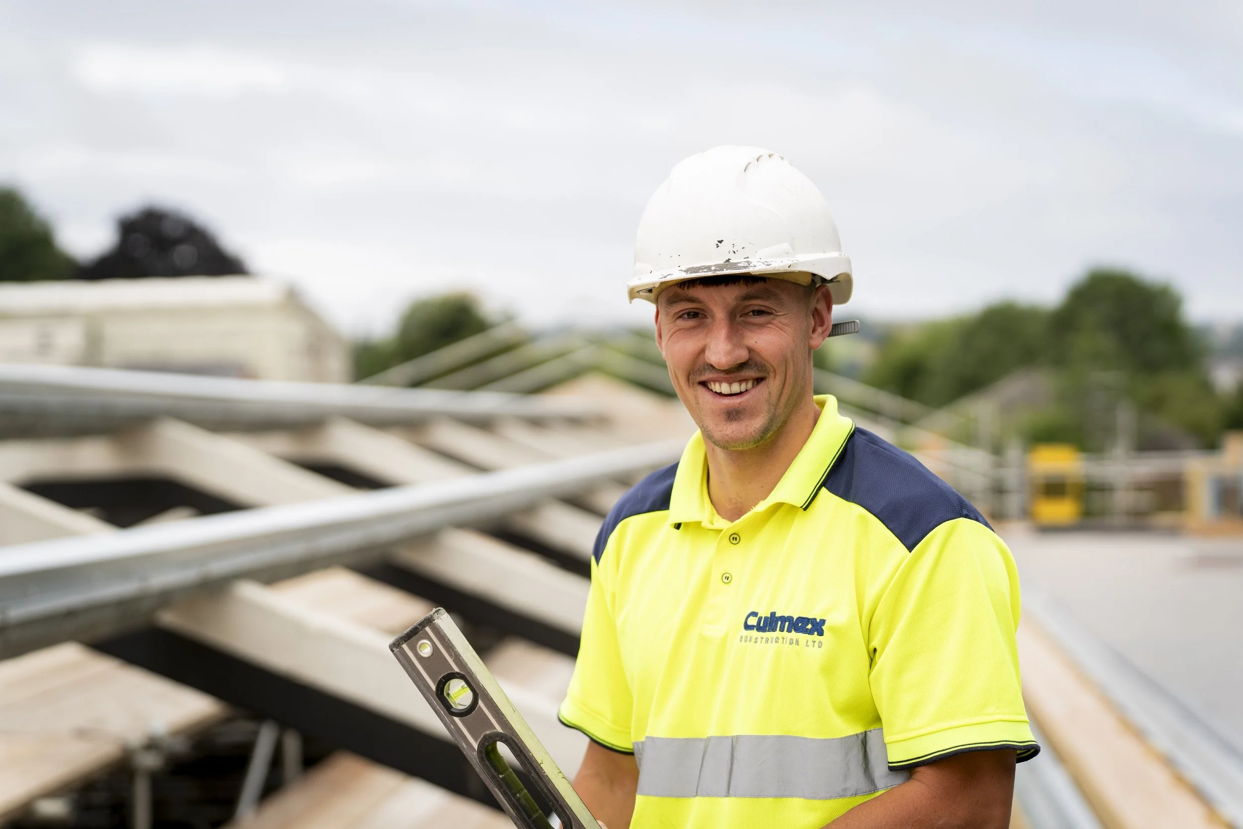 A construction worker wearing a hard hat and high-visibility shirt holds a spirit level on a building site with roof framing behind him. The image is an example of construction photography showing tradespeople and materials.