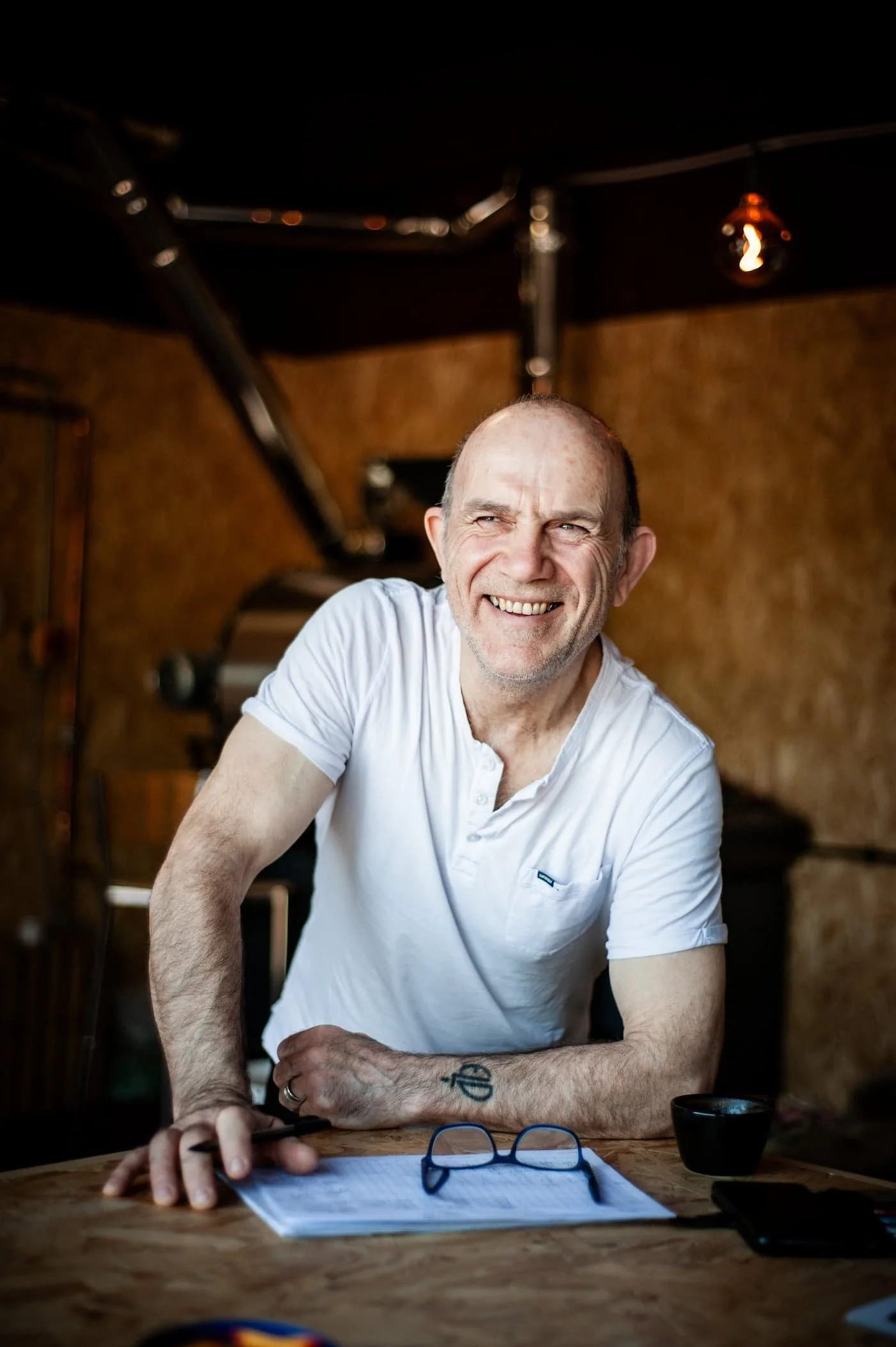 Business portrait photography of a middle-aged white man wearing a white t-shirt. He is sitting at a wooden table which has documents laid out on top of it. The photo was taken by business photographer James Aitken.