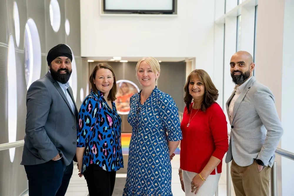Business portrait of five employees standing in a modern office complex. Theya re wearing smart casual business attire and are smiling in a relaxed manner.