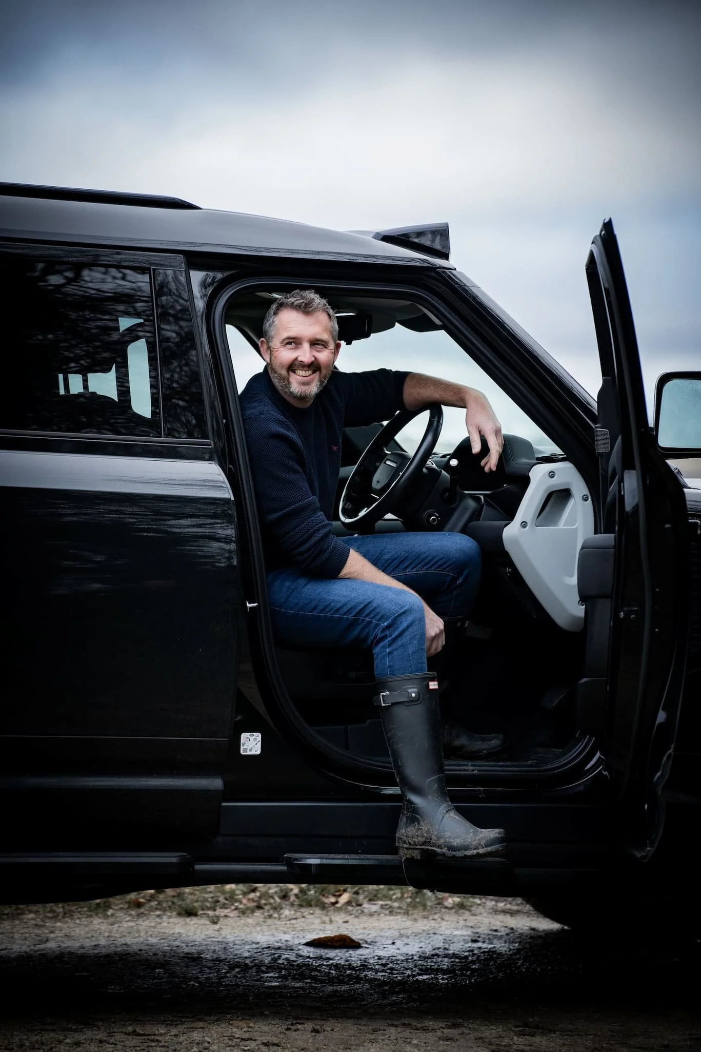 Lifestyle photography of a man sitting in the driver's seat of a black Land Rover Defender. The door is open and he is stepping out of the car.