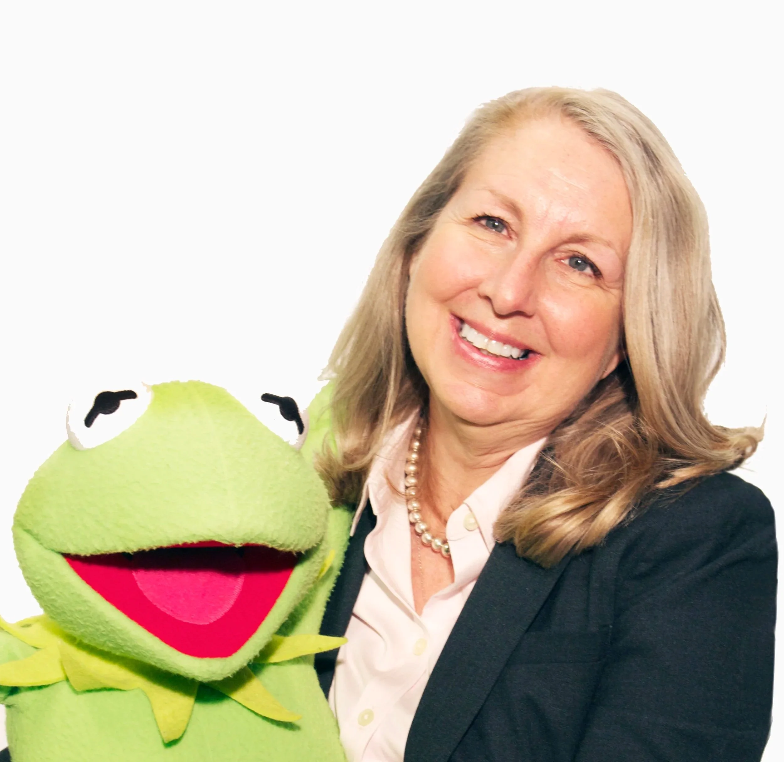 A smiling woman in a black blazer and light pink blouse poses with Kermit the Frog puppet against a white background.