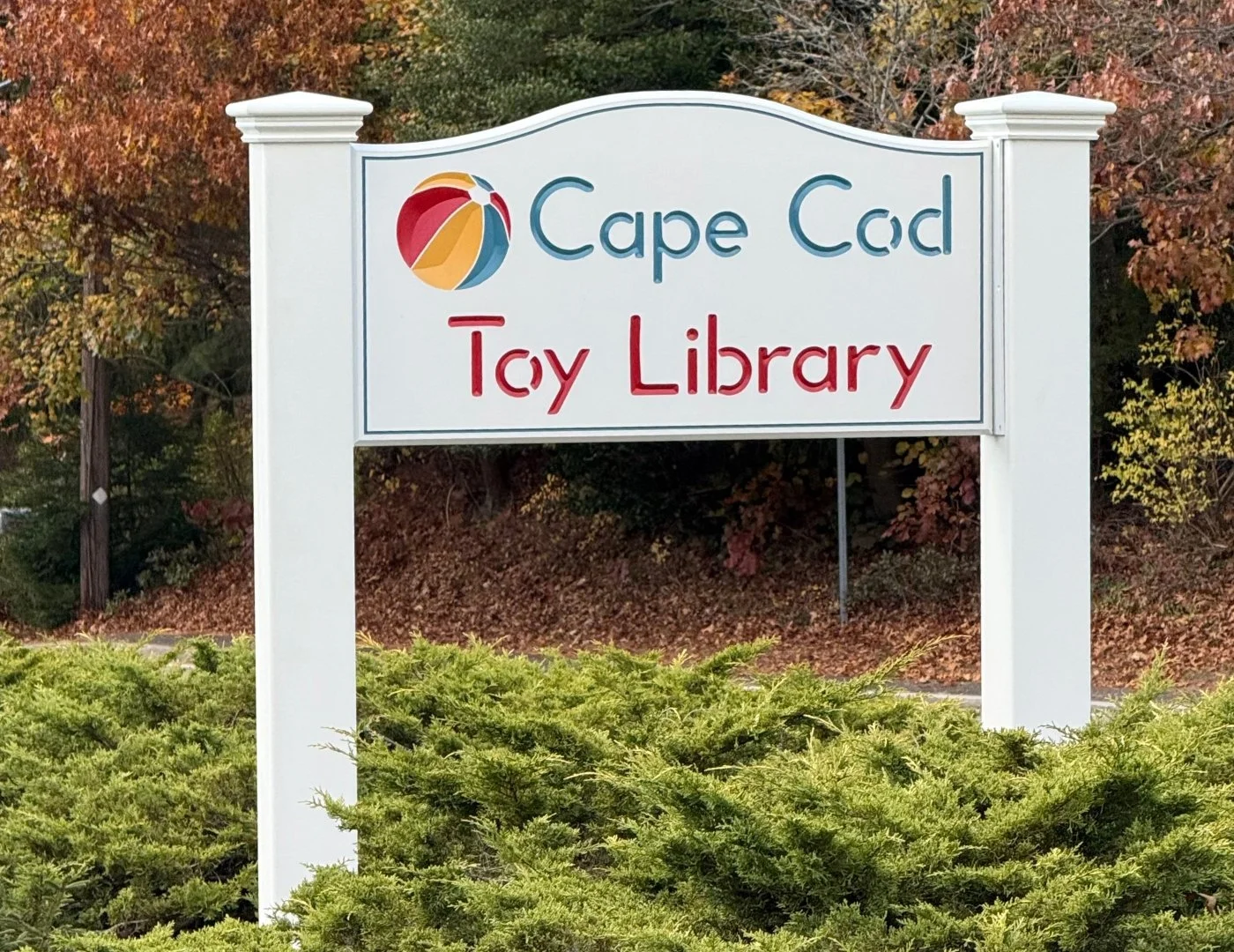 Sign for Cape Cod Toy Library with a colorful beach ball graphic, surrounded by greenery and autumn trees.