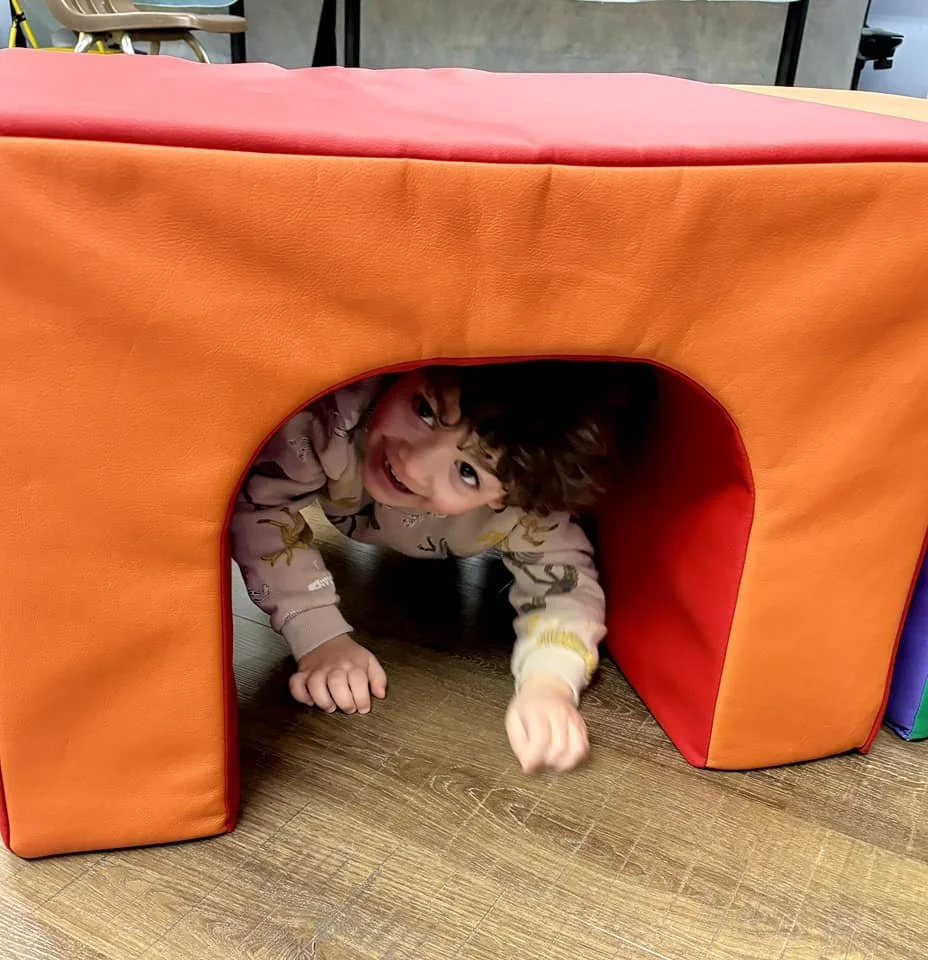 Child crawling through a soft, orange and red play tunnel, smiling and looking at the camera.