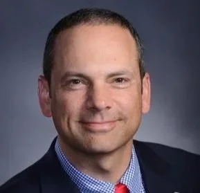 Professional portrait of a man with short dark hair, wearing a dark suit, light blue shirt, and red tie, against a dark gray background.