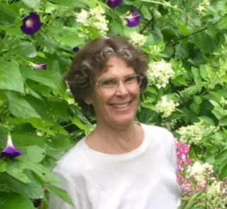 A woman with glasses and short curly hair smiling, standing outdoors among green foliage and purple and white flowers.
