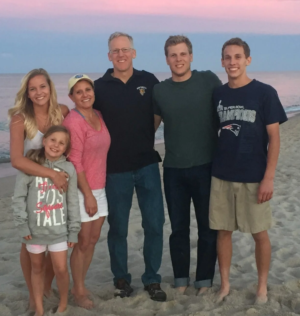 Family of six standing on a beach during sunset, smiling, with ocean and colorful sky in the background.