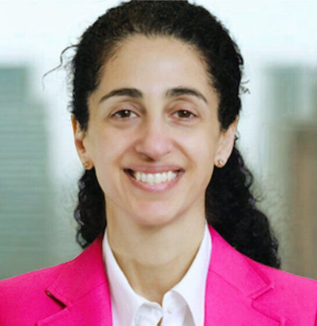 Professional woman with dark curly hair smiling, wearing a pink blazer and white shirt.
