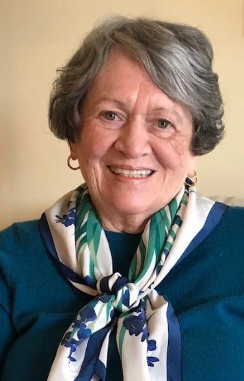A smiling elderly woman with short gray hair, wearing a blue top and a colorful scarf around her neck.