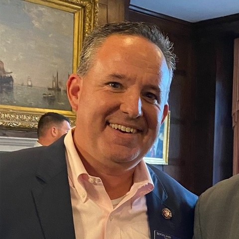 Smiling man in a suit at an indoor event with framed paintings on the wall behind him.