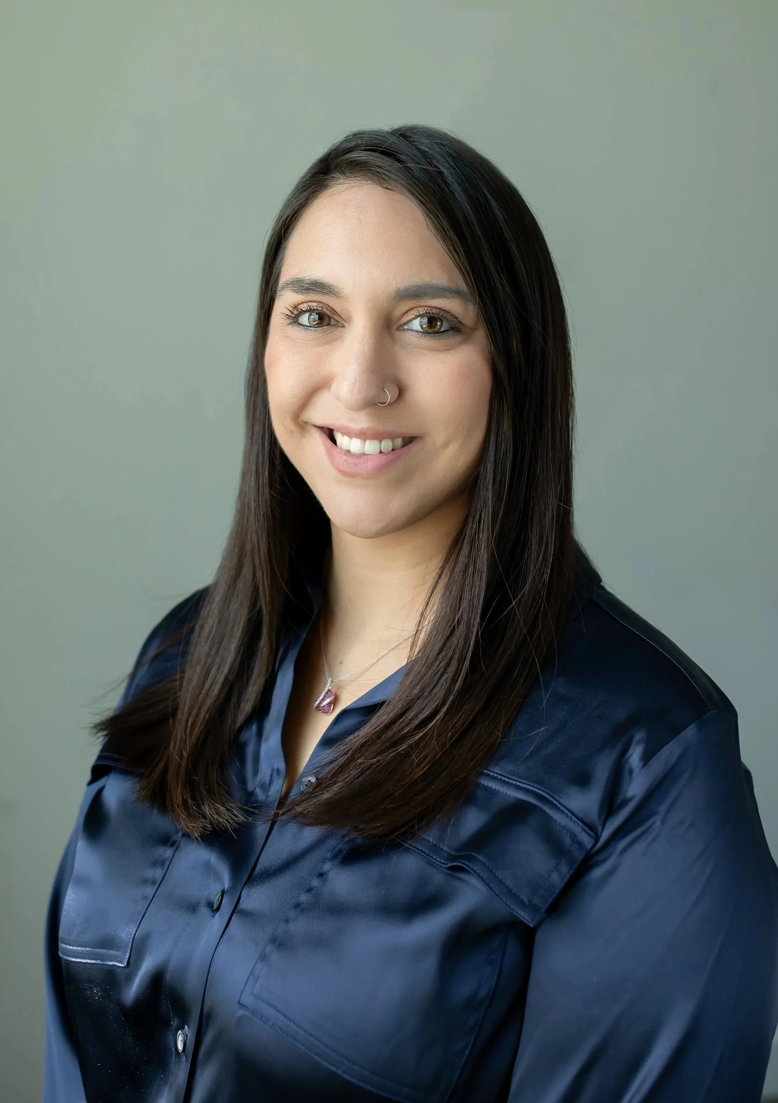 Portrait of a woman with long dark hair, wearing a navy blue satin blouse and a necklace with a red pendant, smiling at the camera against a neutral background.