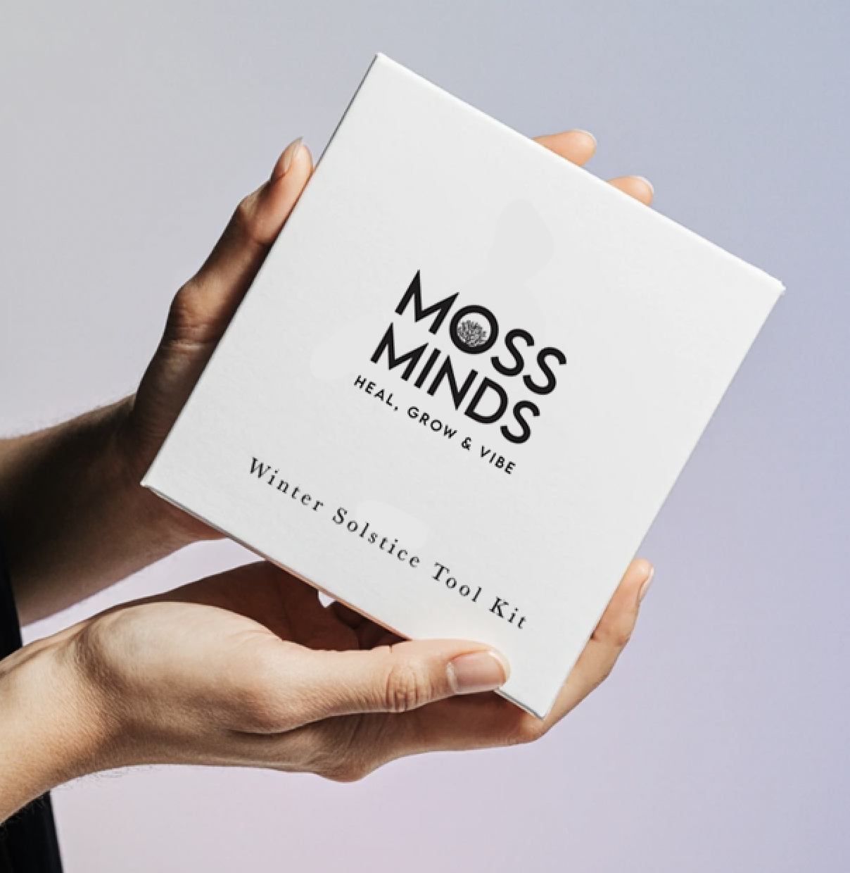 Person holding a box labeled "Moss Minds: Heal, Grow & Vibe - Winter Solstice Tool Kit" against a plain light background.
