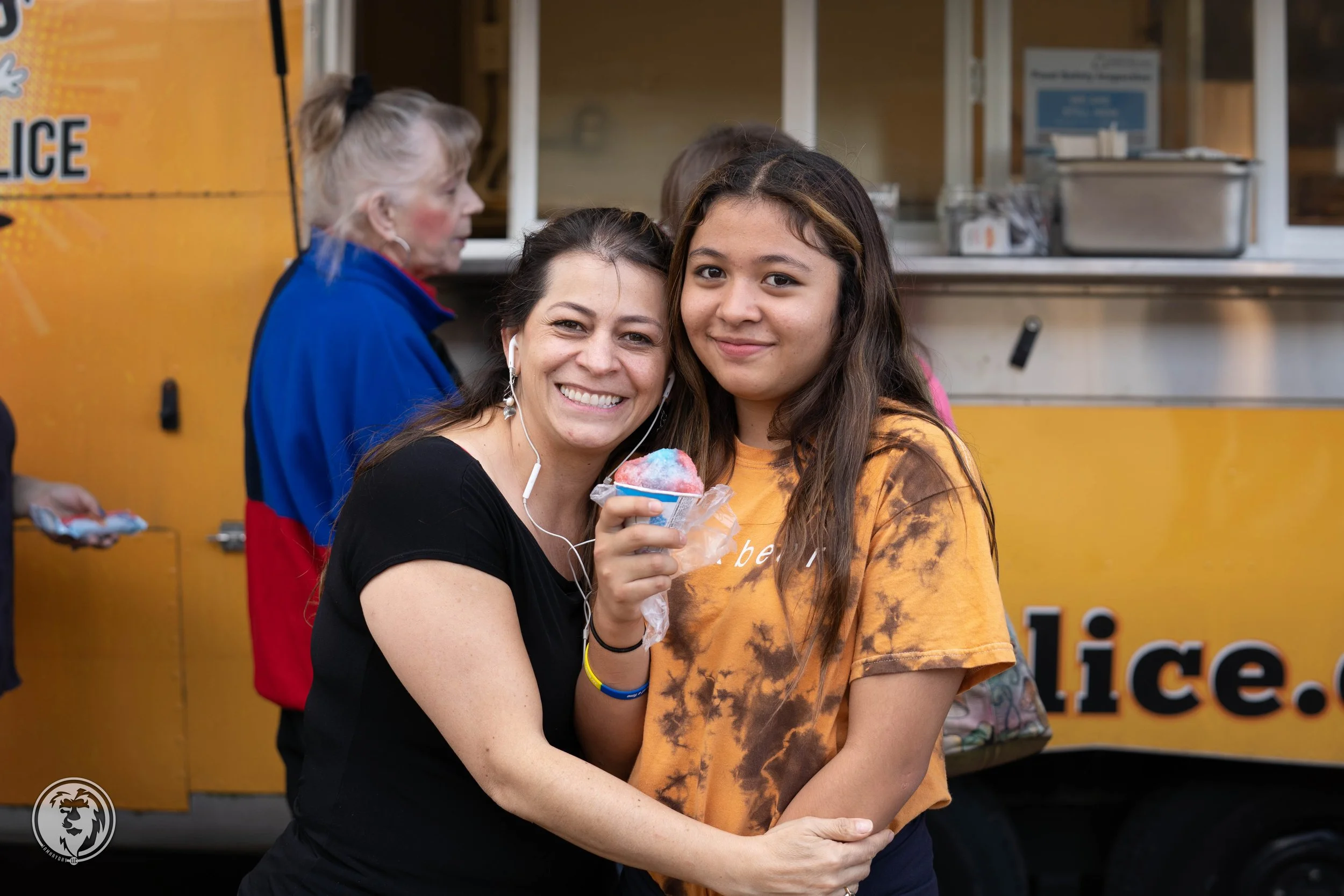 Two people stand in front of a yellow food truck at an outdoor event, smiling.