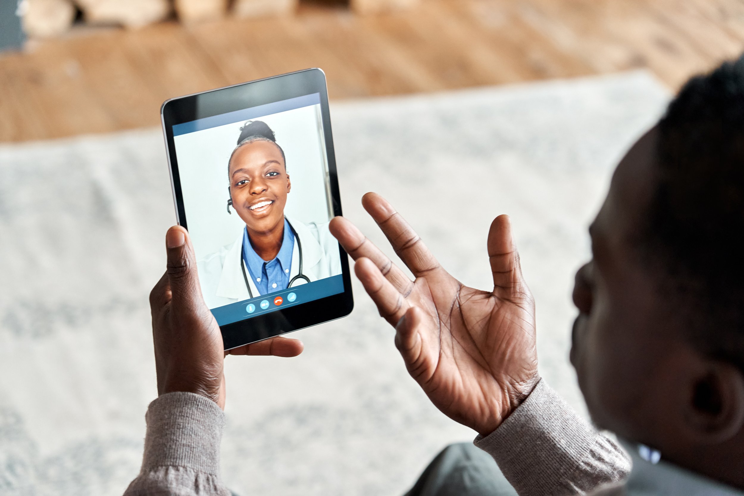 A Black man hold a small tablet on his left hand as he motions with his right hand to imply talking to his doctor (Female/Black) seen on the tablet.