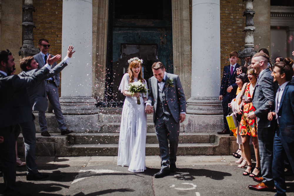 The couple leave the wedding venue as guests through confetti.