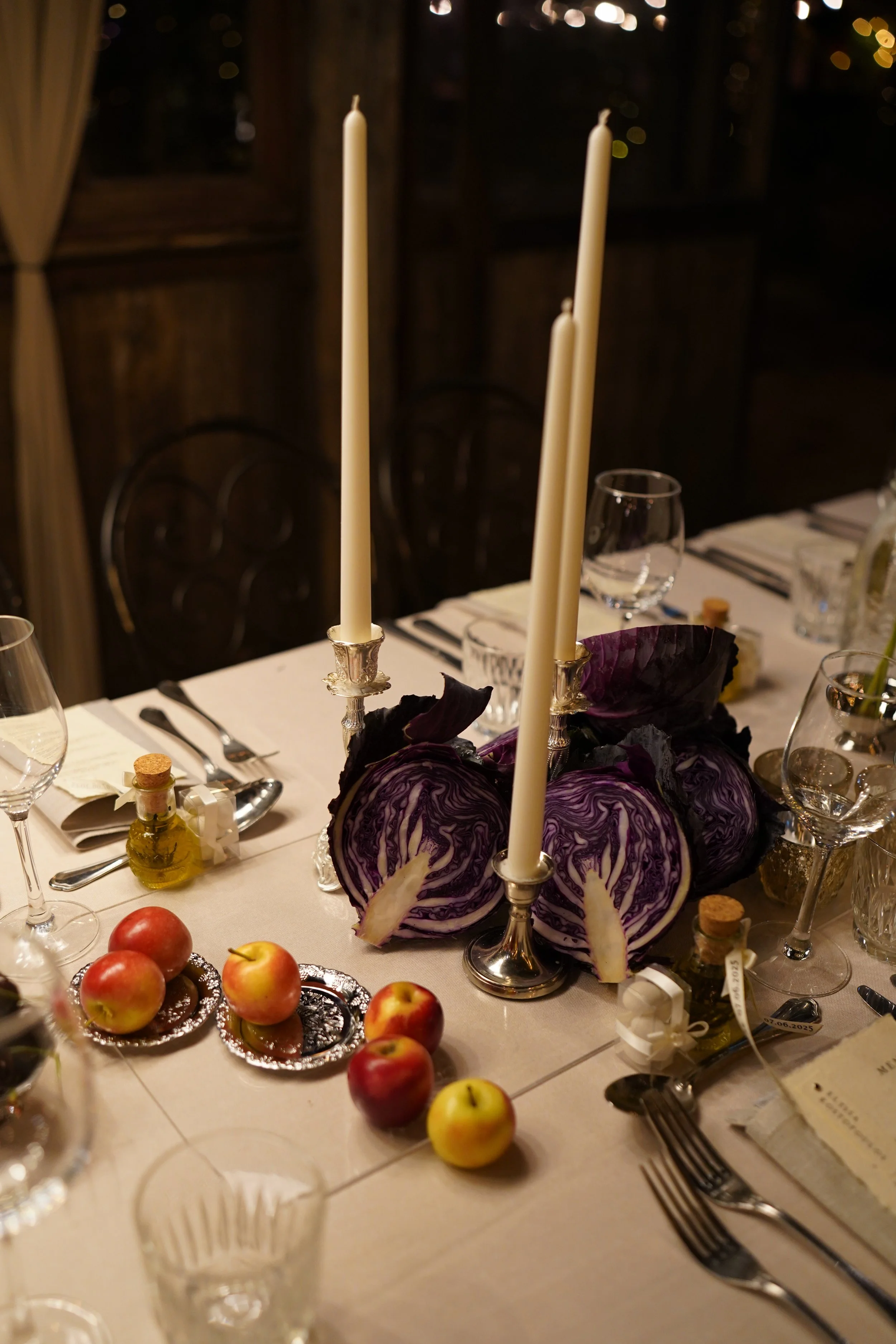 A table decorated with red and green apples, a purple cabbage centerpiece, candles, and glassware for a meal.