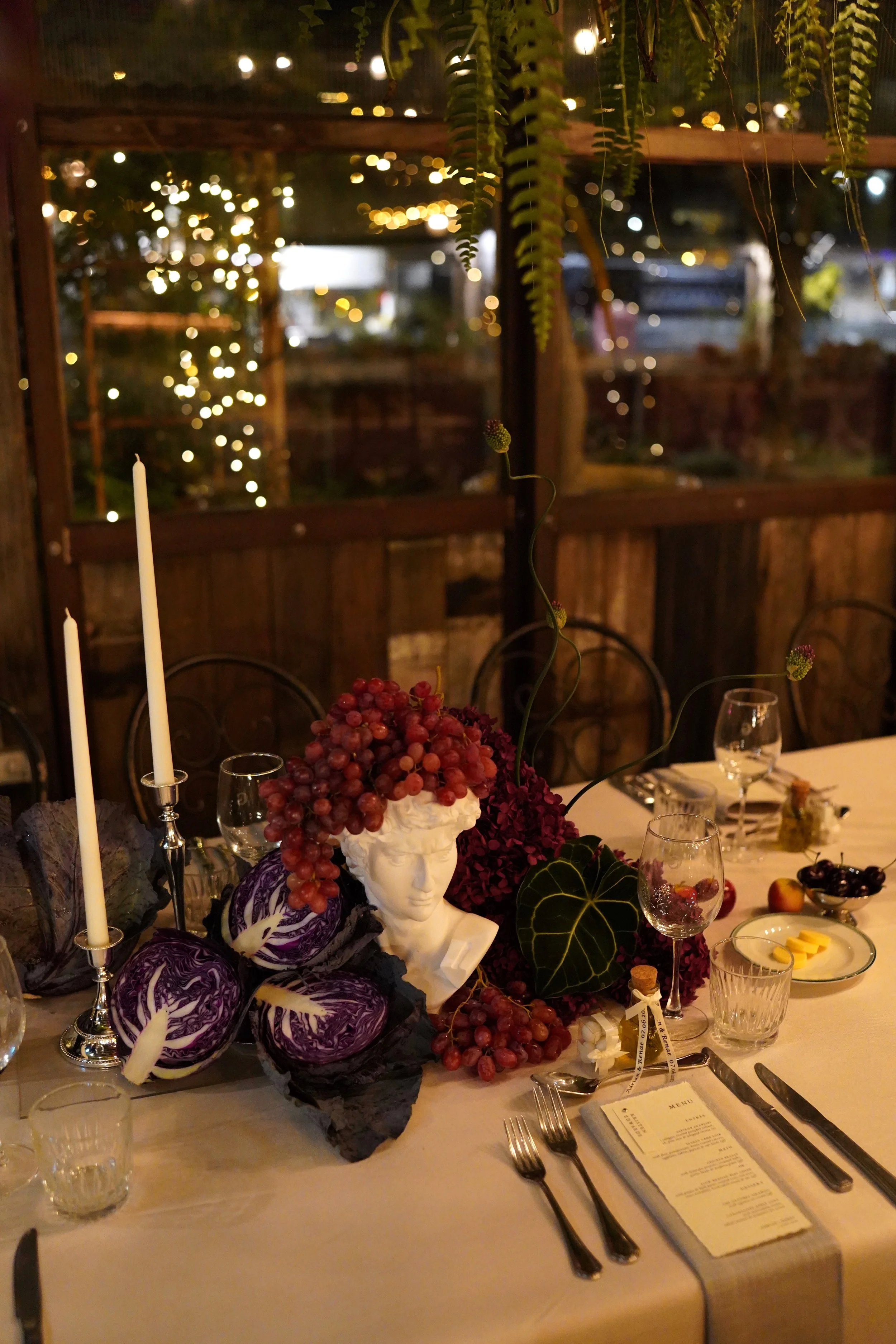 Elegantly set dining table with candles, a grape-themed centerpiece featuring a white bust sculpture, and decorative purple and green vegetables. In the background, a window shows blurred outdoor lights at night.