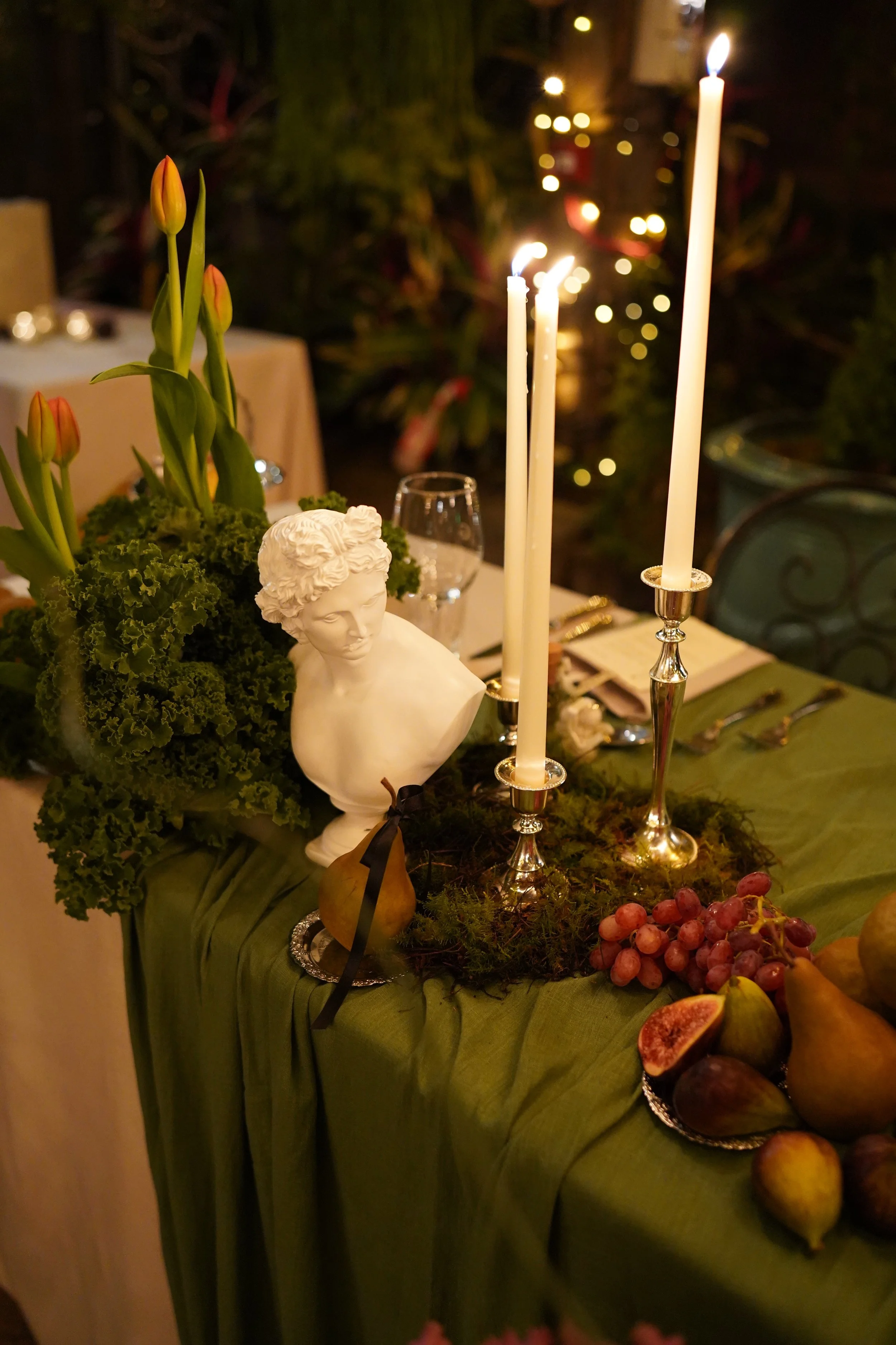 A table centerpiece with a small white bust sculpture, tall white candles in silver holders, a bouquet of tulips with orange and pink buds, grapes, figs, and other fruits, all on a green cloth table cover with blurred warm lights in the background.