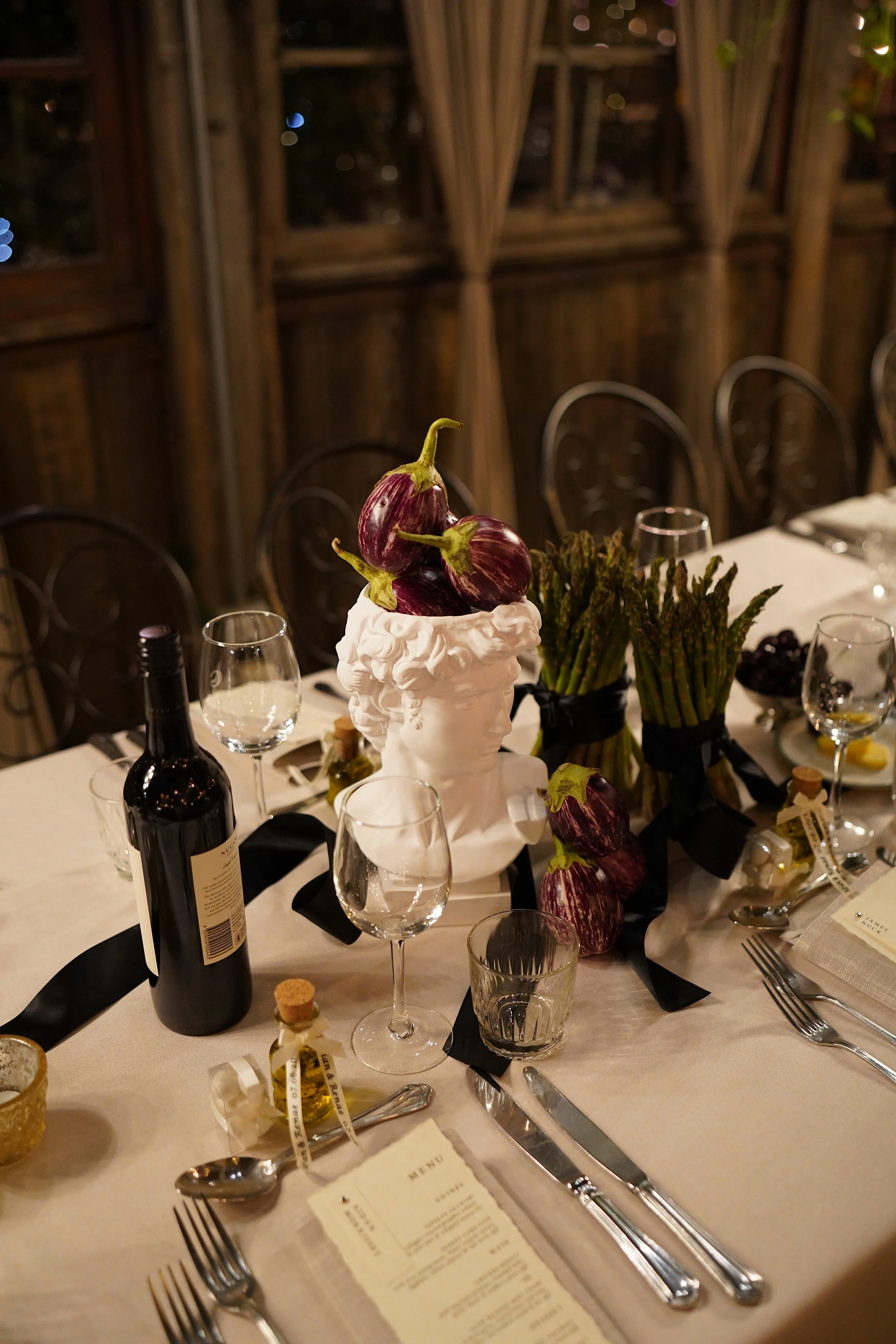 Elegant dinner table setup with wine glasses, a bottle of red wine, a menu, and a centerpiece with eggplants and asparagus at a rustic wooden venue.