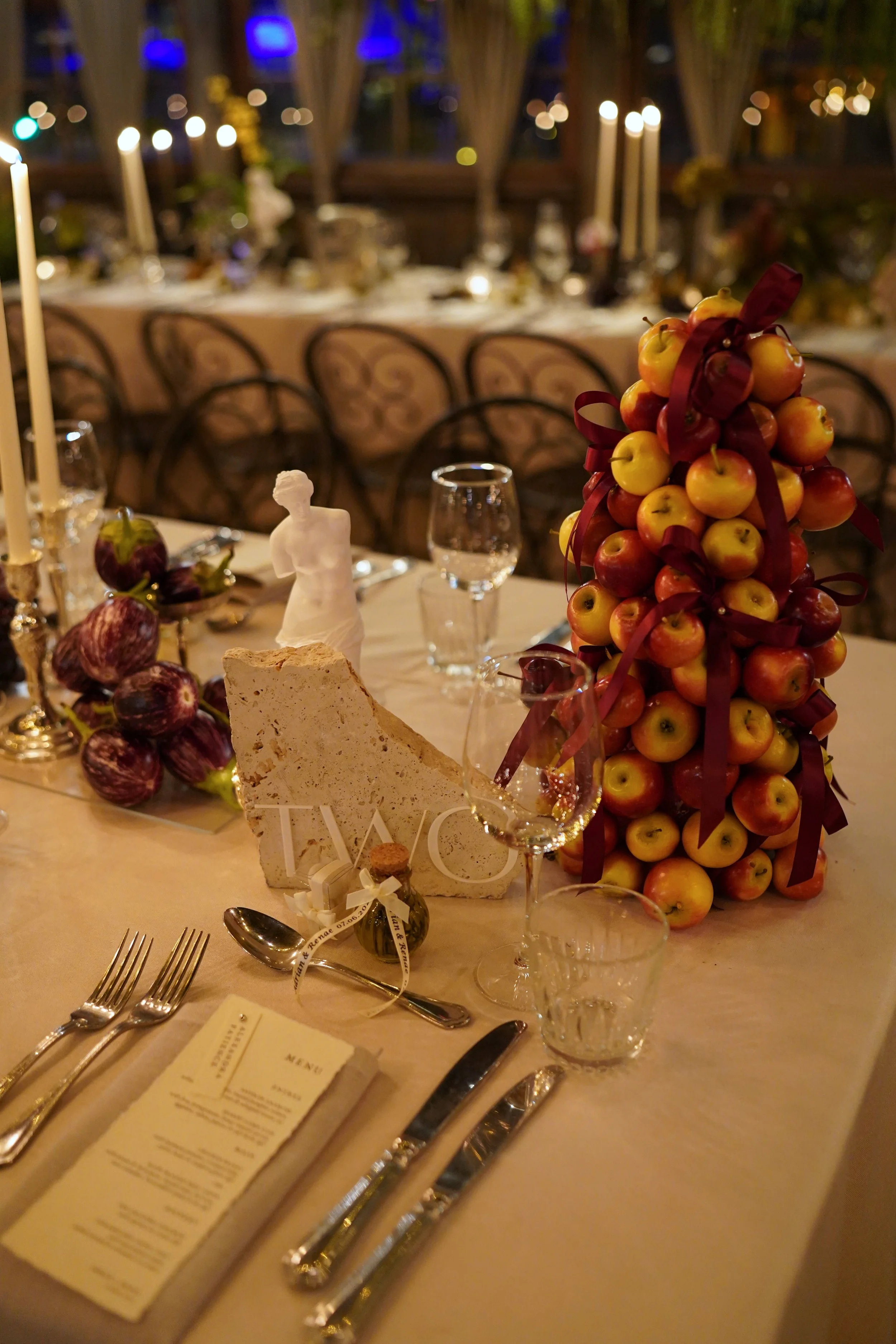 A formal dinner table decorated with a large apple centerpiece tied with a red ribbon, surrounded by apples and figs, with a menu, cutlery, wine glasses, and a small statue of a woman on a beige tablecloth. In the background, there are candles and ch