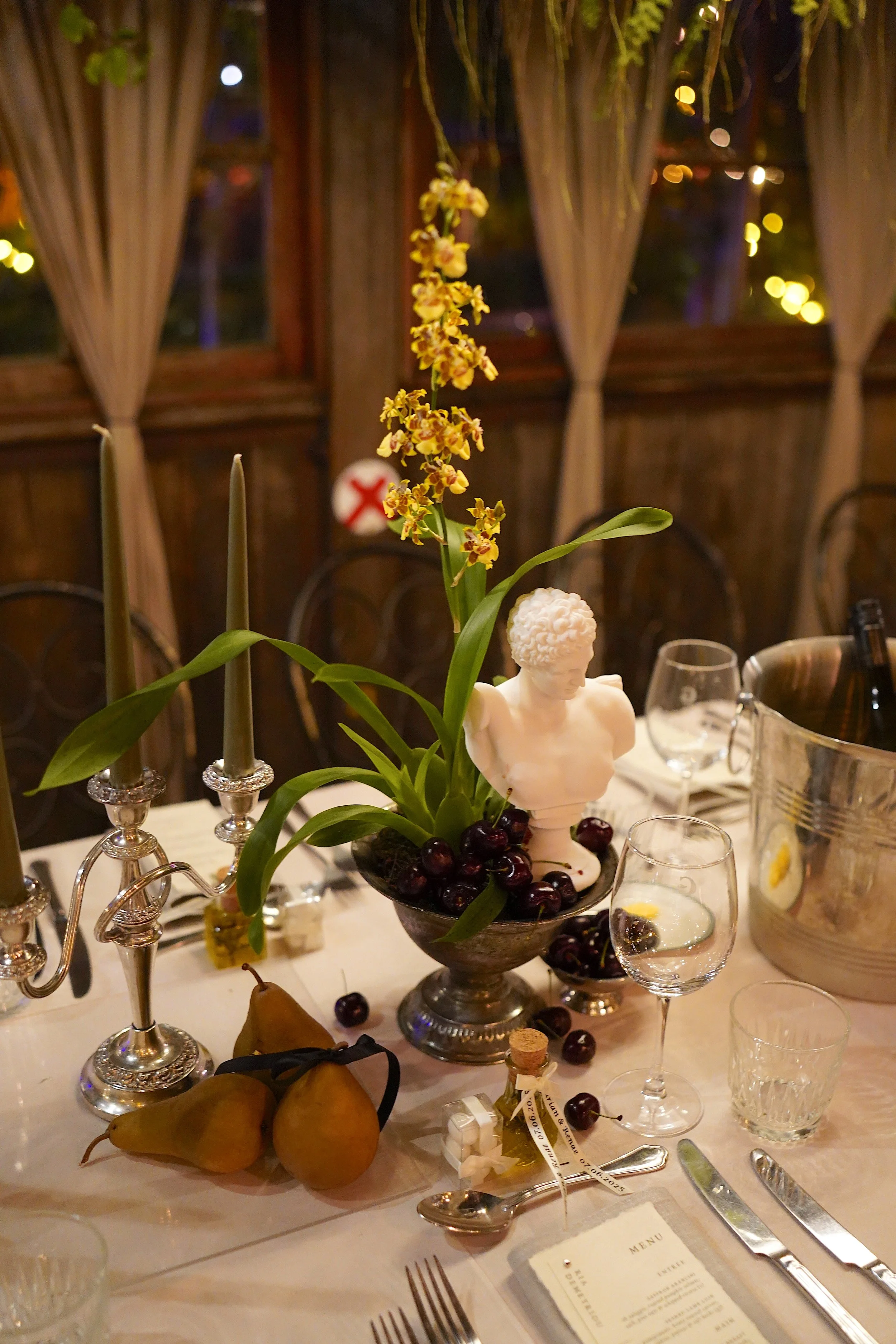 A beautifully set dinner table with a floral centerpiece featuring yellow orchids and a small bust sculpture. The table includes candles, glasses, silverware, and plates. Pears decorated with ribbons are also on the table. In the background, there ar