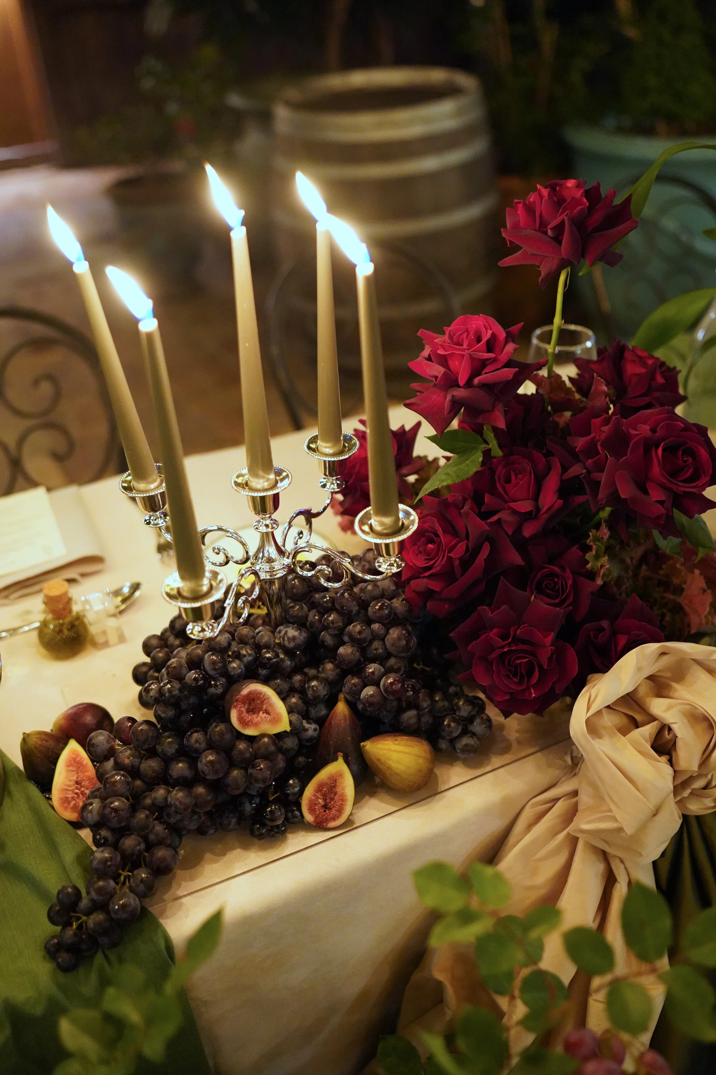 A table decorated with a silver candelabra holding five lit candles, a bunch of dark purple grapes with some sliced open revealing their red insides, fresh figs, and other fruits, surrounded by flowers and greenery.