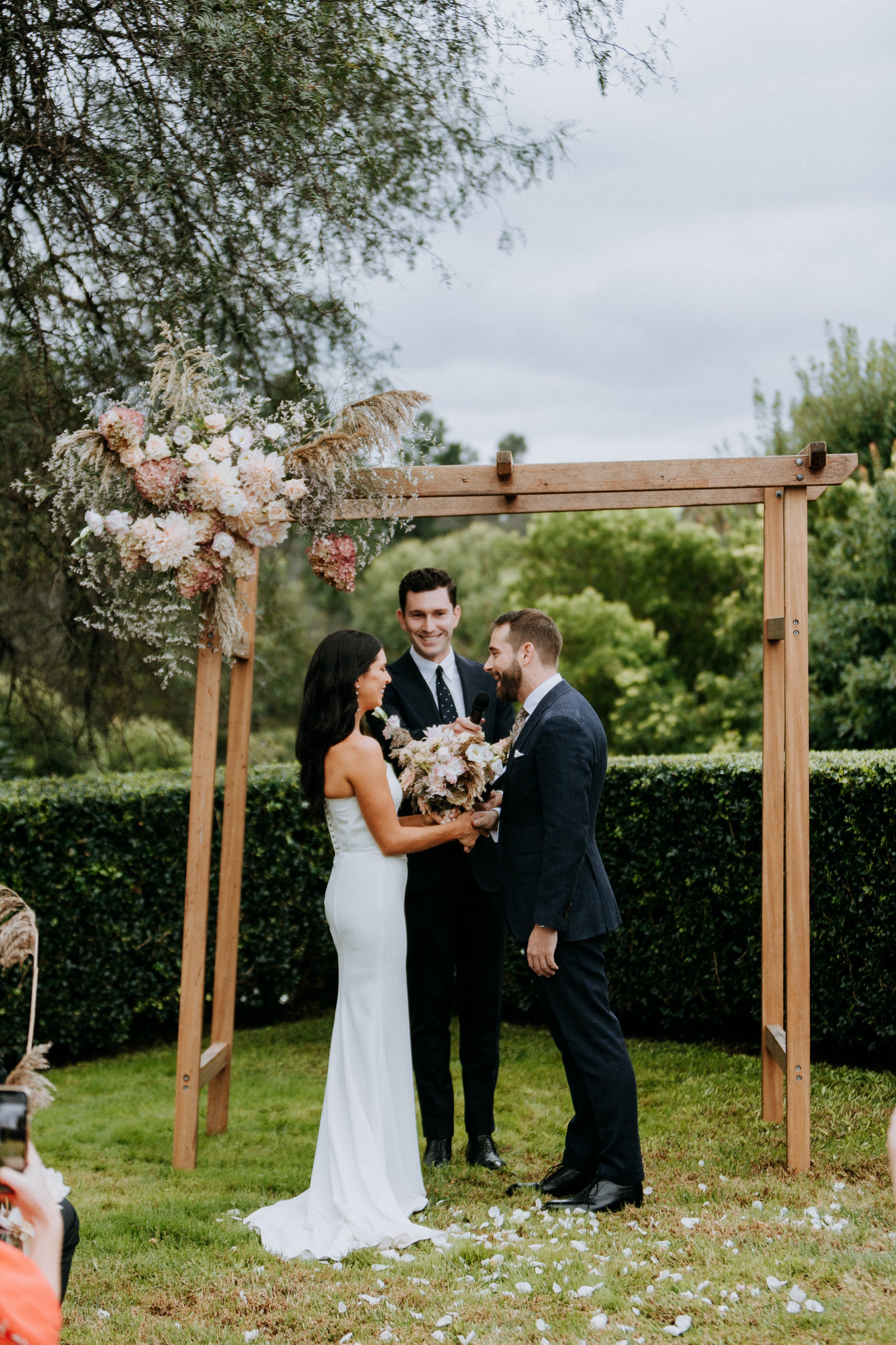 A couple gets married outdoors under a wooden arch decorated with pink and white flowers, with an officiant standing behind them and friends taking photos.
