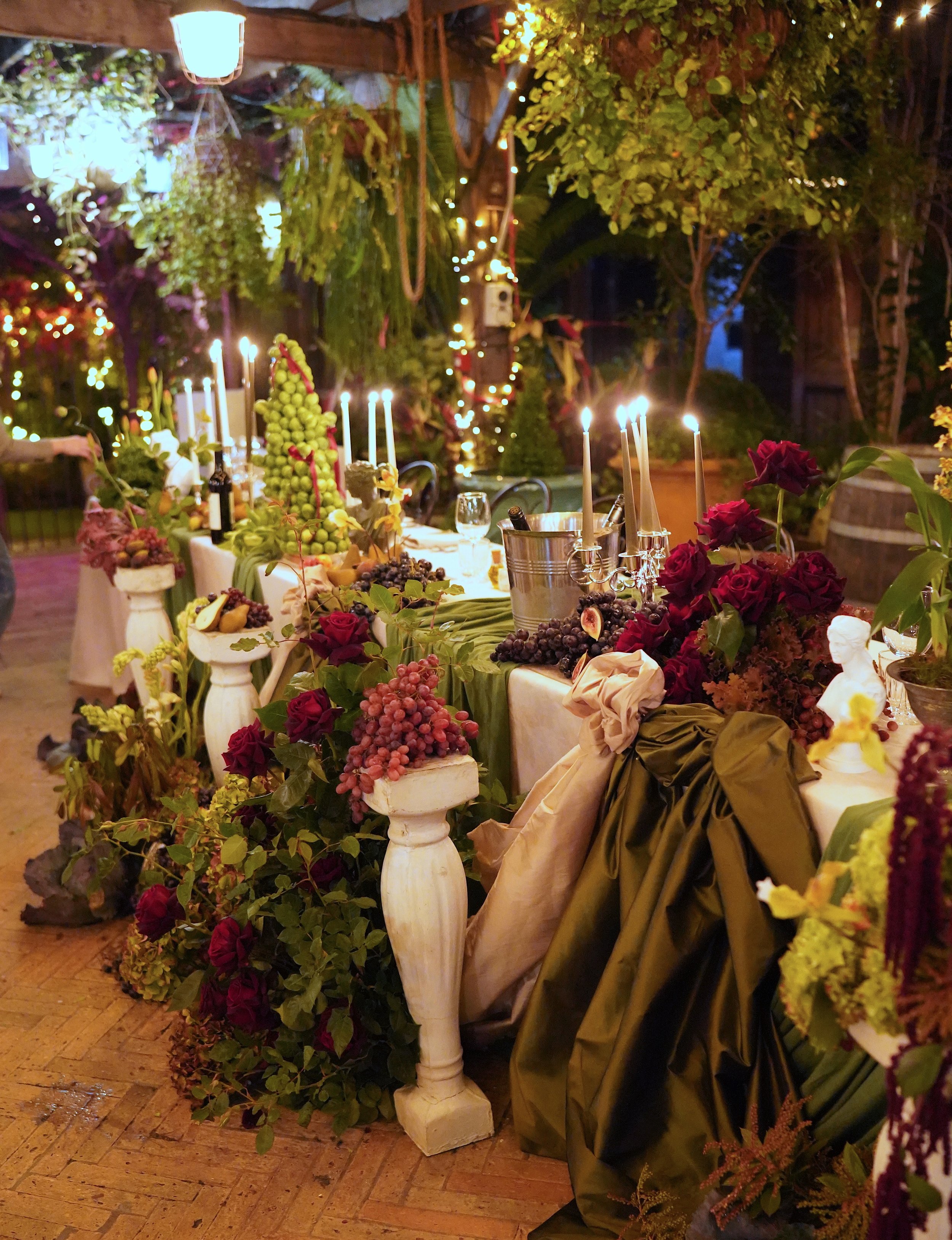 Decorative table setting with fruits, flowers, candles, and greenery at an outdoor evening event, illuminated by string lights and a hanging lamp.